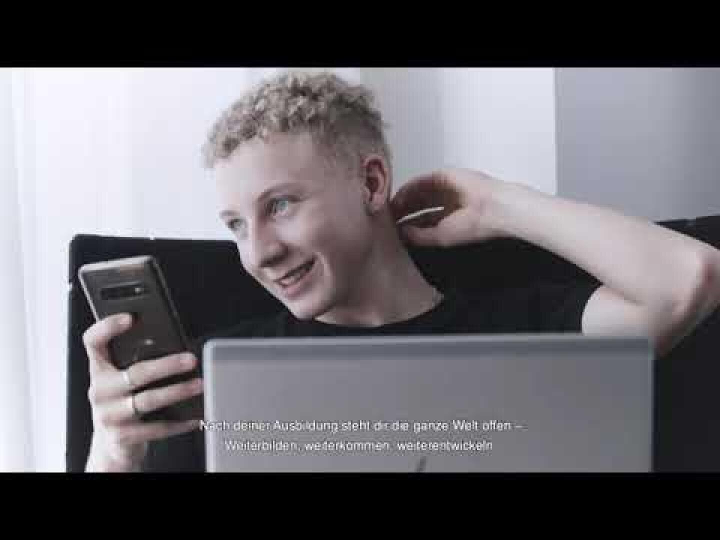 Ein junger Mensch mit lockigem blondem Haar lächelt, während er auf ein Smartphone schaut und hinter einem geöffneten Laptop sitzt. Am unteren Bildrand ist weißer Text auf Deutsch zu sehen.