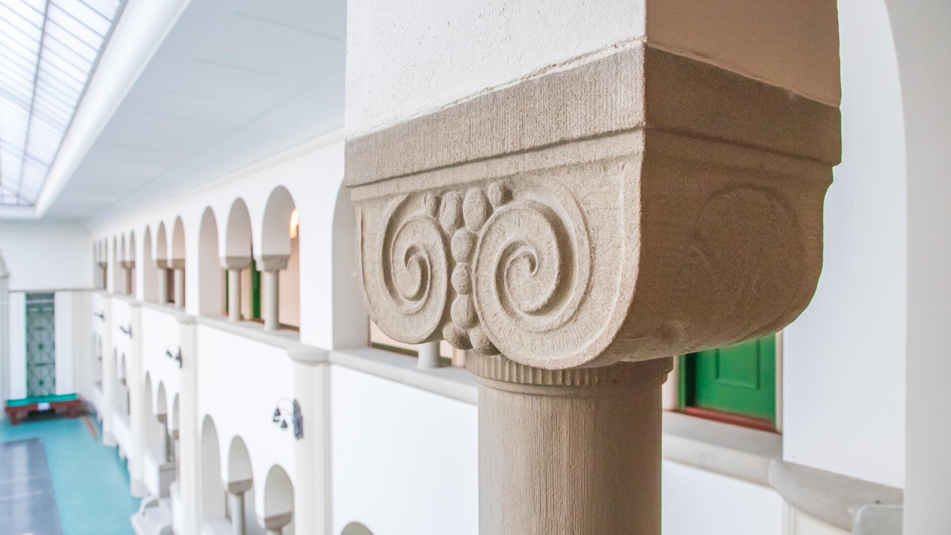 Nahaufnahme einer verschnörkelten Steinsäule mit Spiral- und Blattschnitzereien in einem hellen, von Bögen, weißen Wänden und grünen Fensterläden gesäumten Flur.