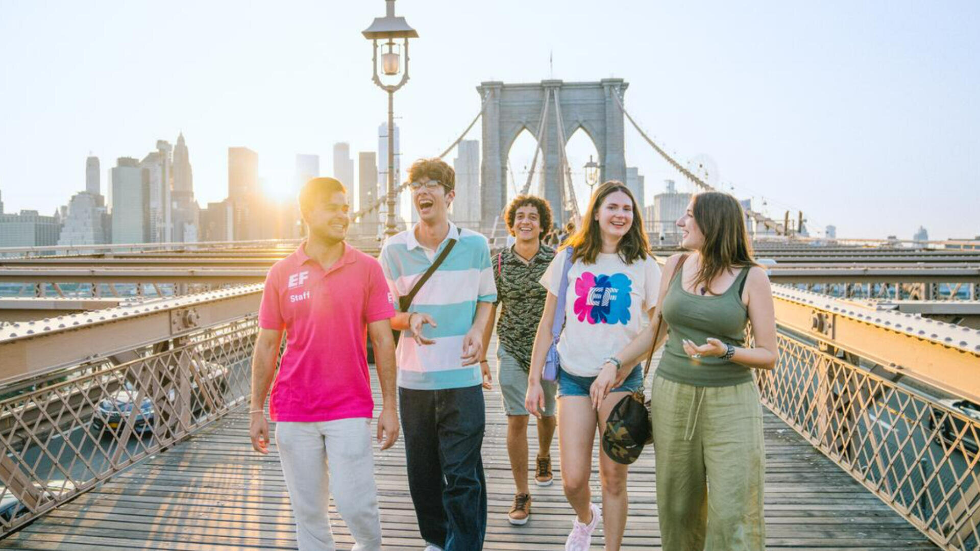 Fünf junge Erwachsene gehen an einem sonnigen Tag auf der Brooklyn Bridge in New York City spazieren und lachen zusammen, während im Hintergrund die Skyline der Stadt zu sehen ist.