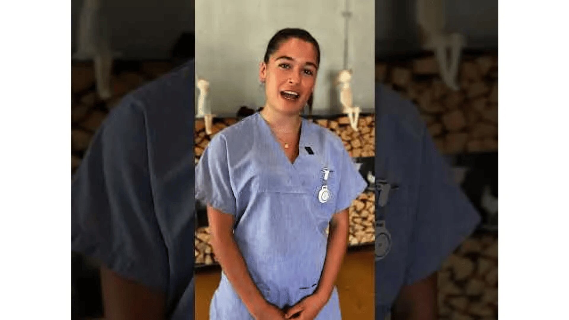 Eine Frau in hellblauem Arztkittel steht in einem Raum, lächelt und spricht. Hinter ihr sind gestapeltes Brennholz und anatomische Modelle in Regalen zu sehen.