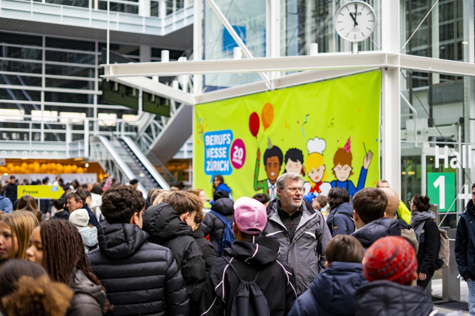 Eine Menschenmenge in Winterkleidung versammelt sich in einem Innenraum in der Nähe eines bunten Schildes der Berufsmesse Zürich mit Cartoon-Figuren. Im Hintergrund sind eine große Uhr und Glaswände zu sehen.