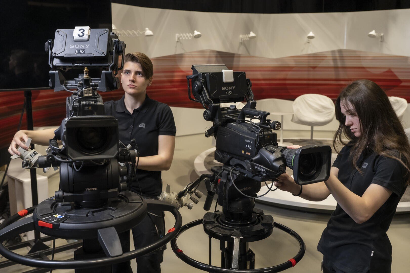 Zwei Frauen in schwarzen Hemden justieren große Videokameras auf Stativen in einem professionellen Studio mit leeren weißen Stühlen und einem roten und beigen abstrakten Hintergrund.