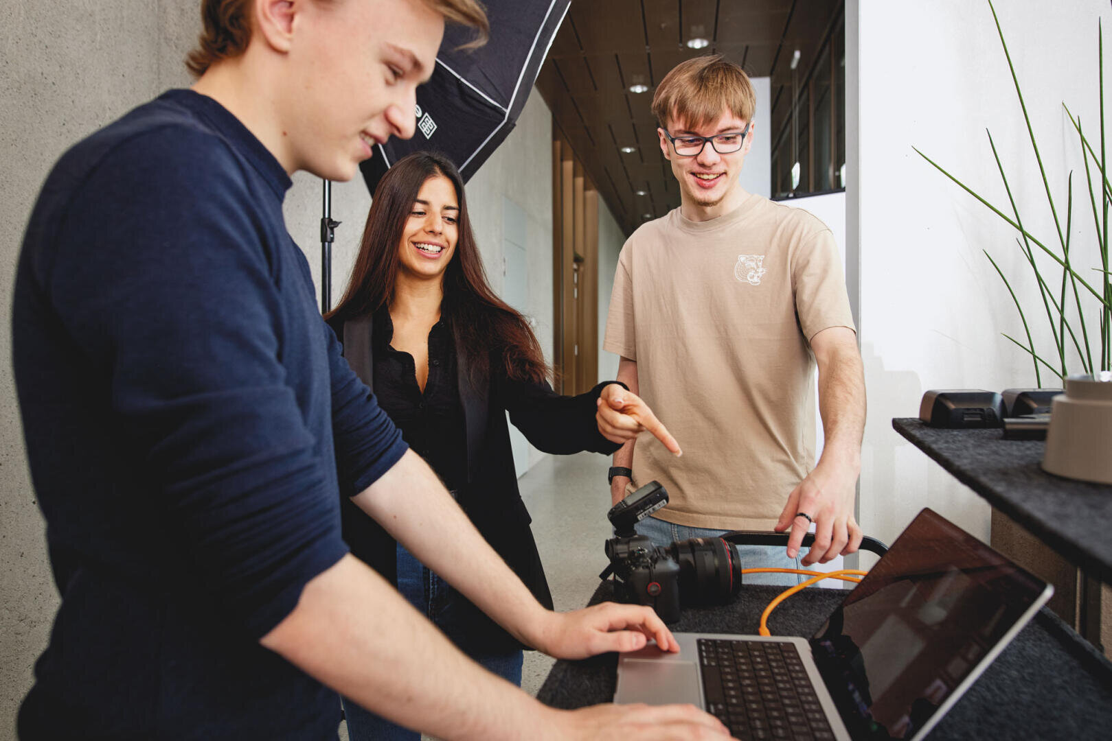 Drei junge Erwachsene arbeiten gemeinsam in einem Raum mit einem Laptop und einer Kameraausrüstung. Eine Person tippt auf dem Laptop, eine andere zeigt auf die Kamera, und eine dritte lächelt, während sie das Geschehen beobachtet.
