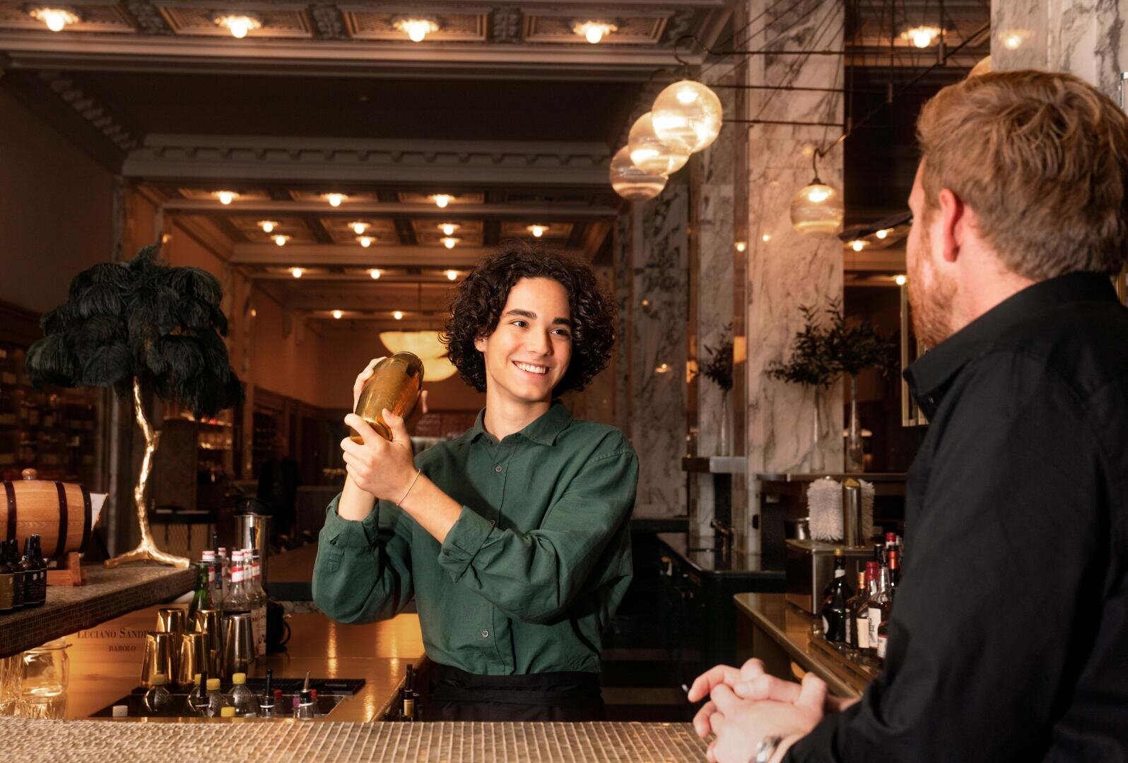 Ein lächelnder Barkeeper in einem grünen Hemd schüttelt hinter einer Marmor-Theke einen Cocktailshaker, während ihm ein schwarz gekleideter Gast gegenüber sitzt – unter warmer, eleganter Beleuchtung in einer stilvollen Baratmosphäre.