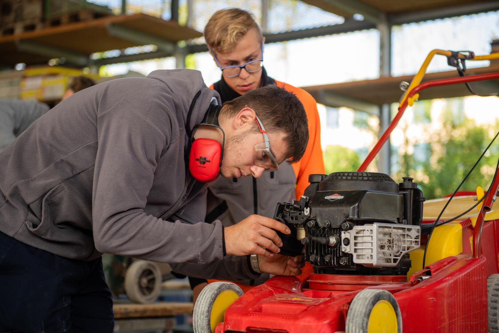 Zwei junge Männer, die Schutzbrillen und Arbeitskleidung tragen, untersuchen und reparieren den Motor eines roten Rasenmähers in einer Werkstatt. Der eine beugt sich nahe heran, um den Motor zu untersuchen, während der andere beobachtet.