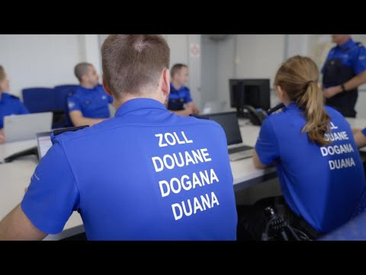 Mehrere Zollbeamte in blauen Uniformen sitzen mit Laptops an einem Tisch. Auf den Rücken zweier Hemden der Beamten steht das Wort „Zoll“ in mehreren Sprachen: Zoll, Douane, Dogana und Duana.
