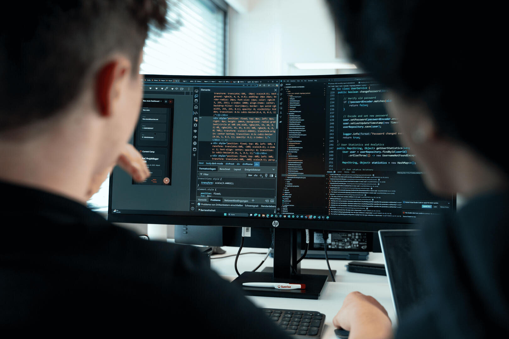 In einer modernen Büroumgebung sitzen zwei Personen an einem Schreibtisch und konzentrieren sich auf zwei Monitore, auf denen Code und Softwareentwicklungswerkzeuge angezeigt werden.