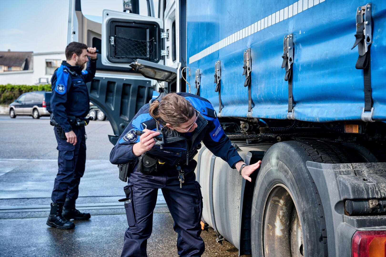 Zwei Polizisten in blauen Uniformen untersuchen einen großen blauen Lastwagen. Einer untersucht den Hinterreifen genau, während der andere mit einem Funkgerät in der Hand neben der offenen Fahrerhaustür steht.