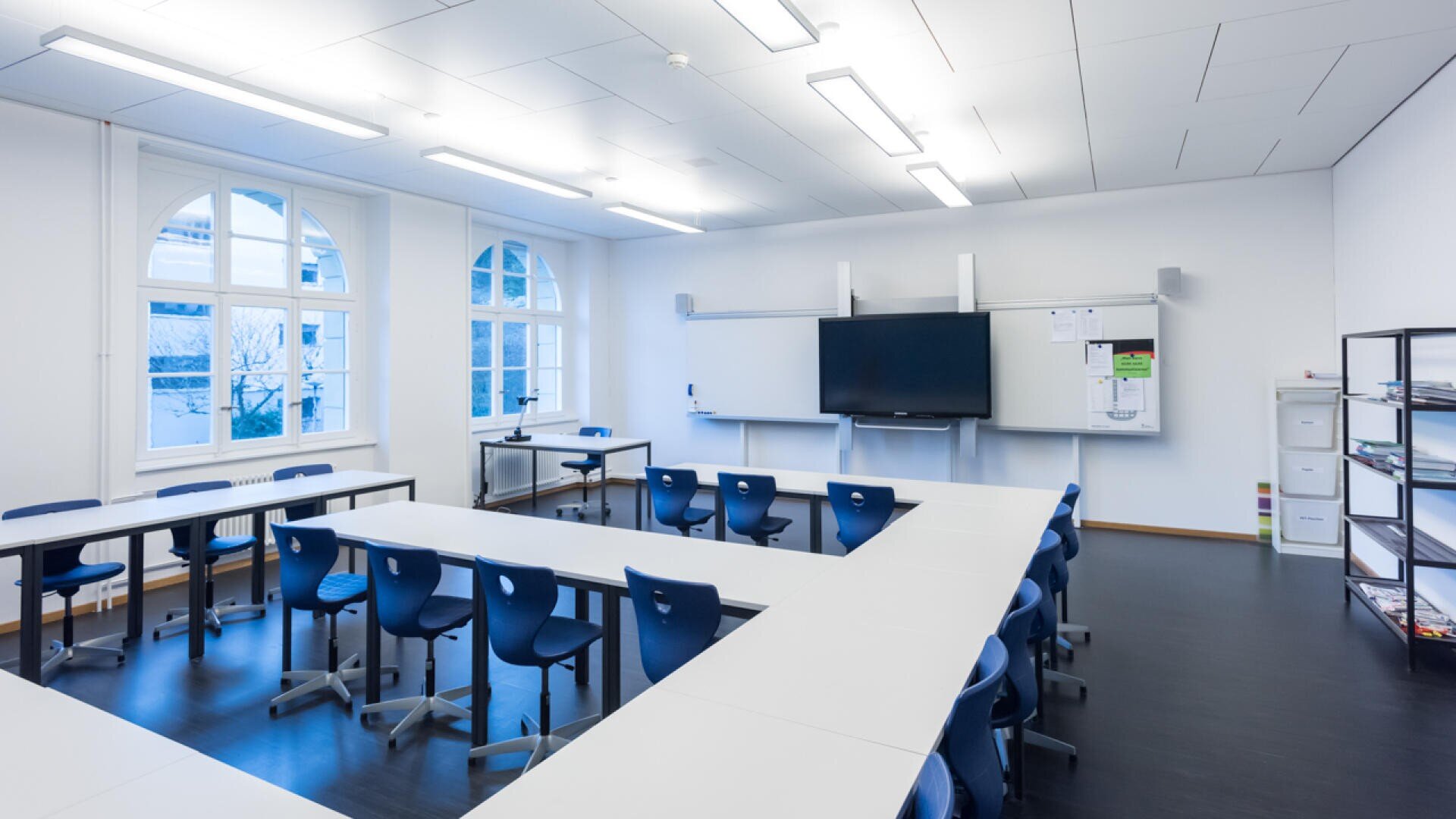 Helles, modernes Klassenzimmer mit blauen Stühlen und weißen Tischen, die in U-Form angeordnet sind, großen Fenstern, einer Tafel, einem an der Wand befestigten Bildschirm und Ablagefächern an der rechten Wand. Der Raum ist leer und gut beleuchtet.