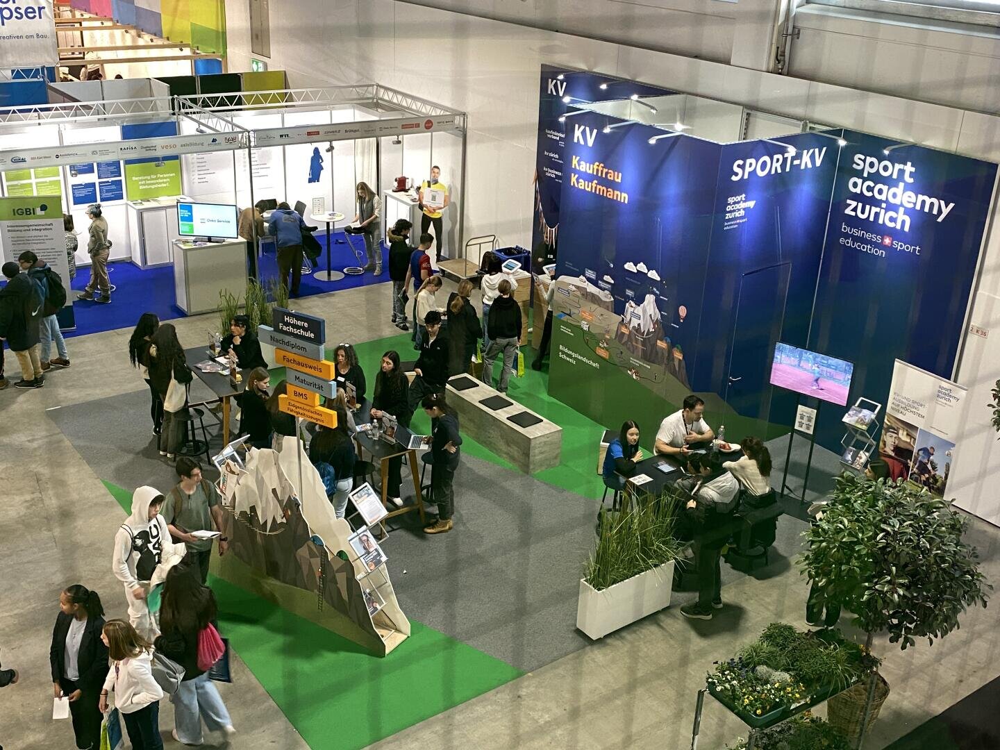 Draufsicht auf eine Indoor-Messe mit Menschen, die Stände besuchen. Der Hauptstand hat blaue Wände mit der Aufschrift SPORT-KV und Sport academy zürich. Die Besucher interagieren mit den Ausstellern und erkunden die Stände im gesamten Raum.