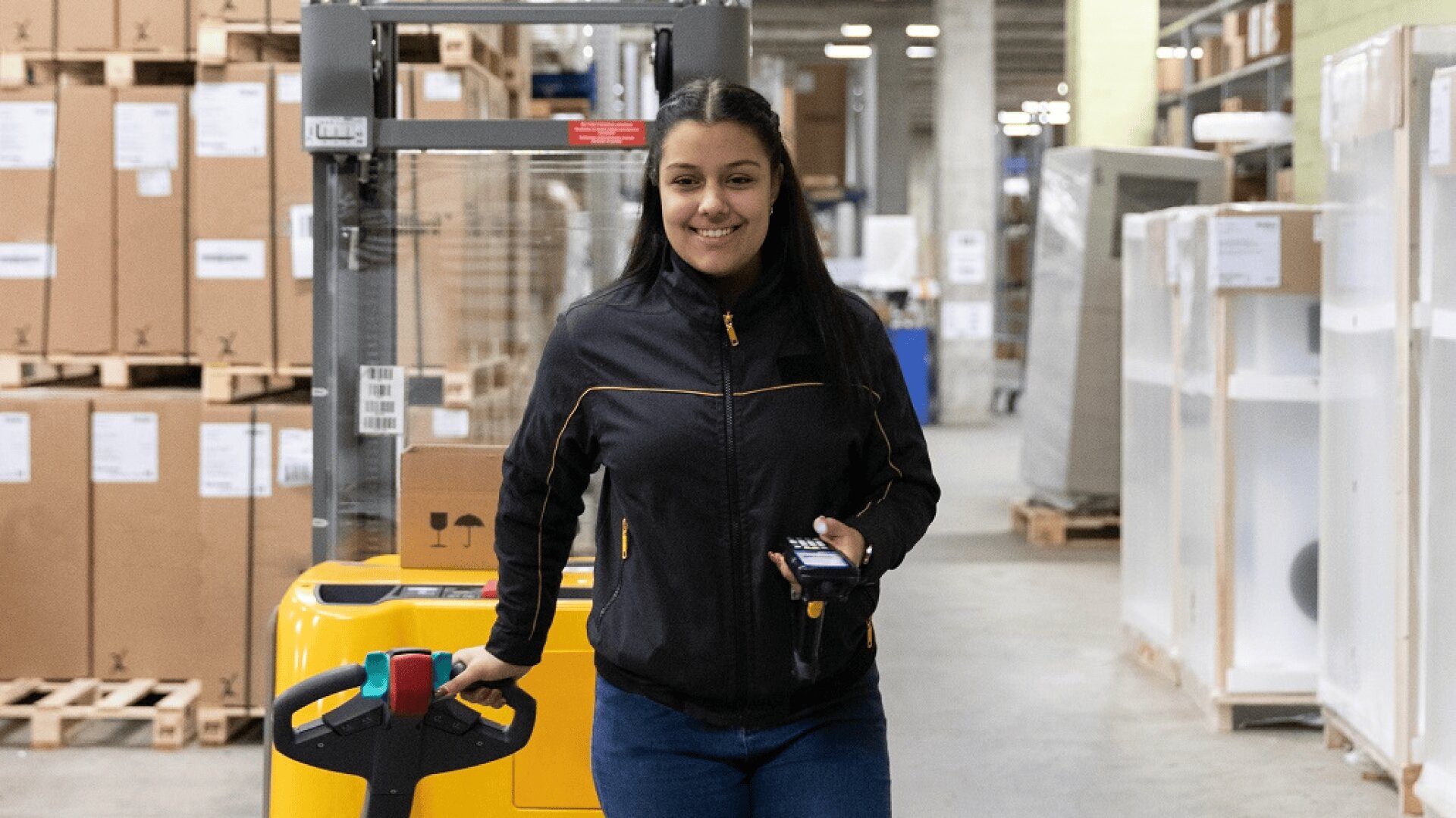 Eine Frau mit einer schwarzen Jacke steht lächelnd in einem Lagerhaus und hält in der einen Hand ein Mobilgerät und in der anderen den Griff eines gelben Hubwagens. Im Hintergrund sind Kisten und Regale zu sehen.