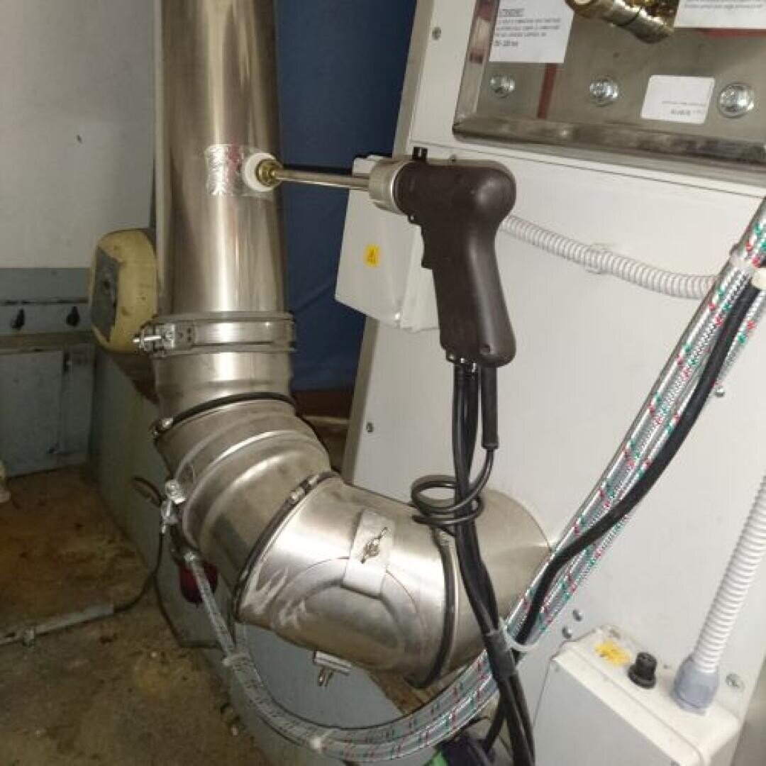 Ein Handgerät mit einem Kabel ist an einem großen Metallrohr in der Nähe eines Bedienfelds befestigt, wahrscheinlich zu Mess- oder Testzwecken in einer industriellen Umgebung.