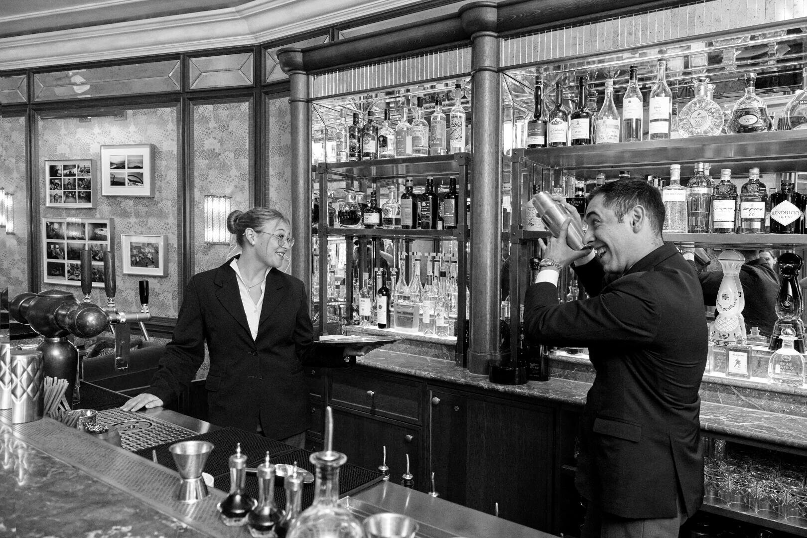 Ein Barkeeper und ein Kellner, beide in formeller Kleidung, unterhalten sich spielerisch hinter einer mit Flaschen und Gläsern bestückten Bar. Der Barkeeper schüttelt einen Cocktail, während der Kellner ihn lächelnd beobachtet. Die Szene ist in schwarz-weiß gehalten.