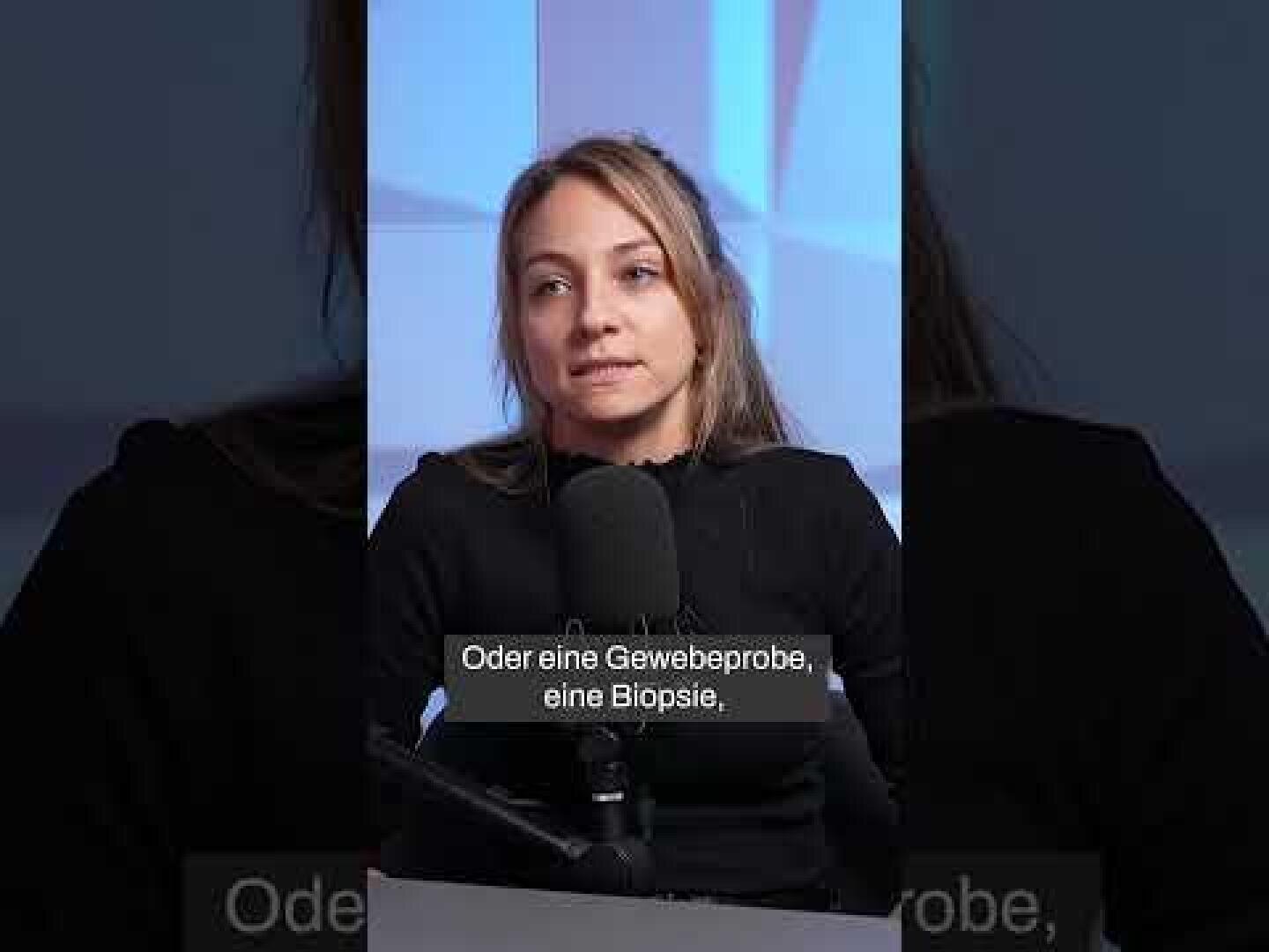 Eine Frau mit langen blonden Haaren, die ein schwarzes Oberteil trägt, spricht in einem Studio in ein Mikrofon. Die deutschen Untertitel am unteren Rand lauten: "Oder eine Gewebeprobe, eine Biopsie".