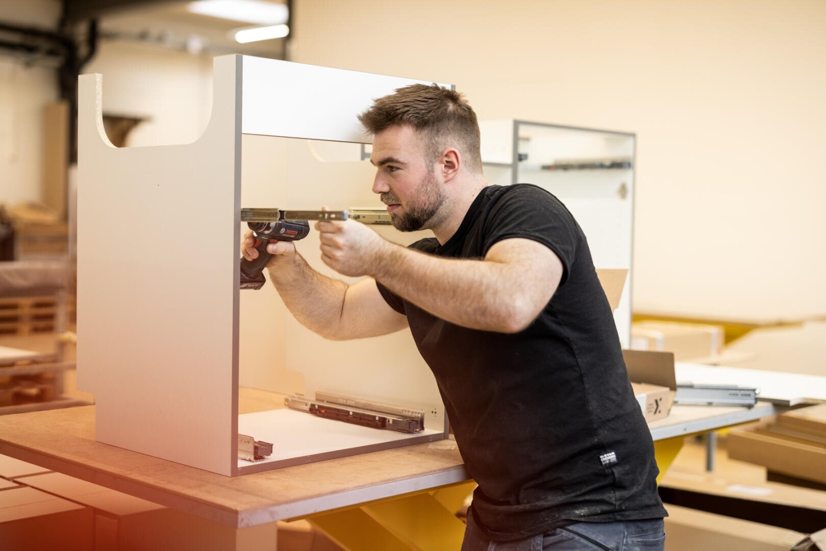 Ein Mann in einem schwarzen T-Shirt baut in einer Werkstatt mit einer Bohrmaschine einen weißen Schrank zusammen und konzentriert sich dabei darauf, eine Metallschiene im Inneren des Schranks zu befestigen.