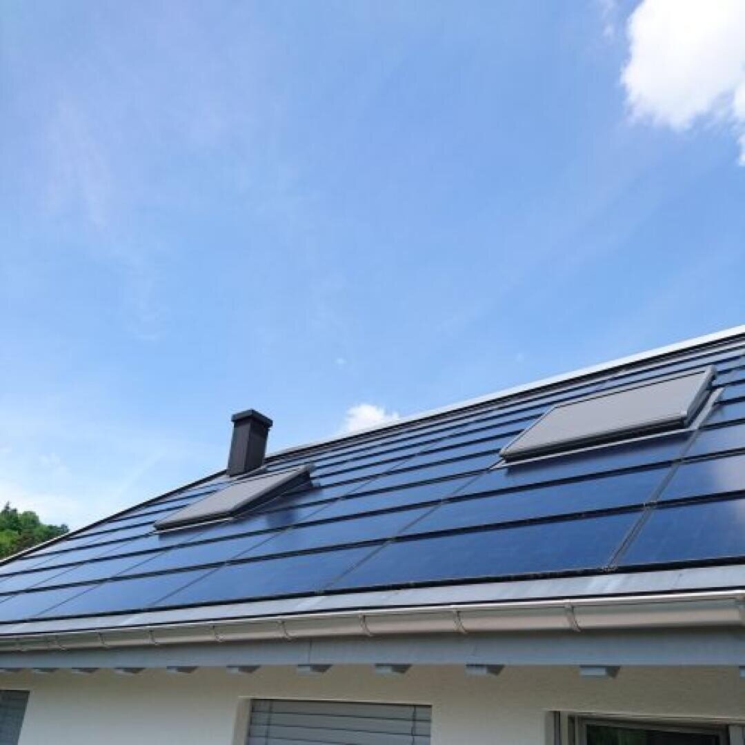 Ein schräges Dach mit Sonnenkollektoren und zwei Oberlichtern, unter einem blauen Himmel mit einigen weißen Wolken. Die weiße Außenwand des Hauses ist im unteren Bereich teilweise sichtbar.