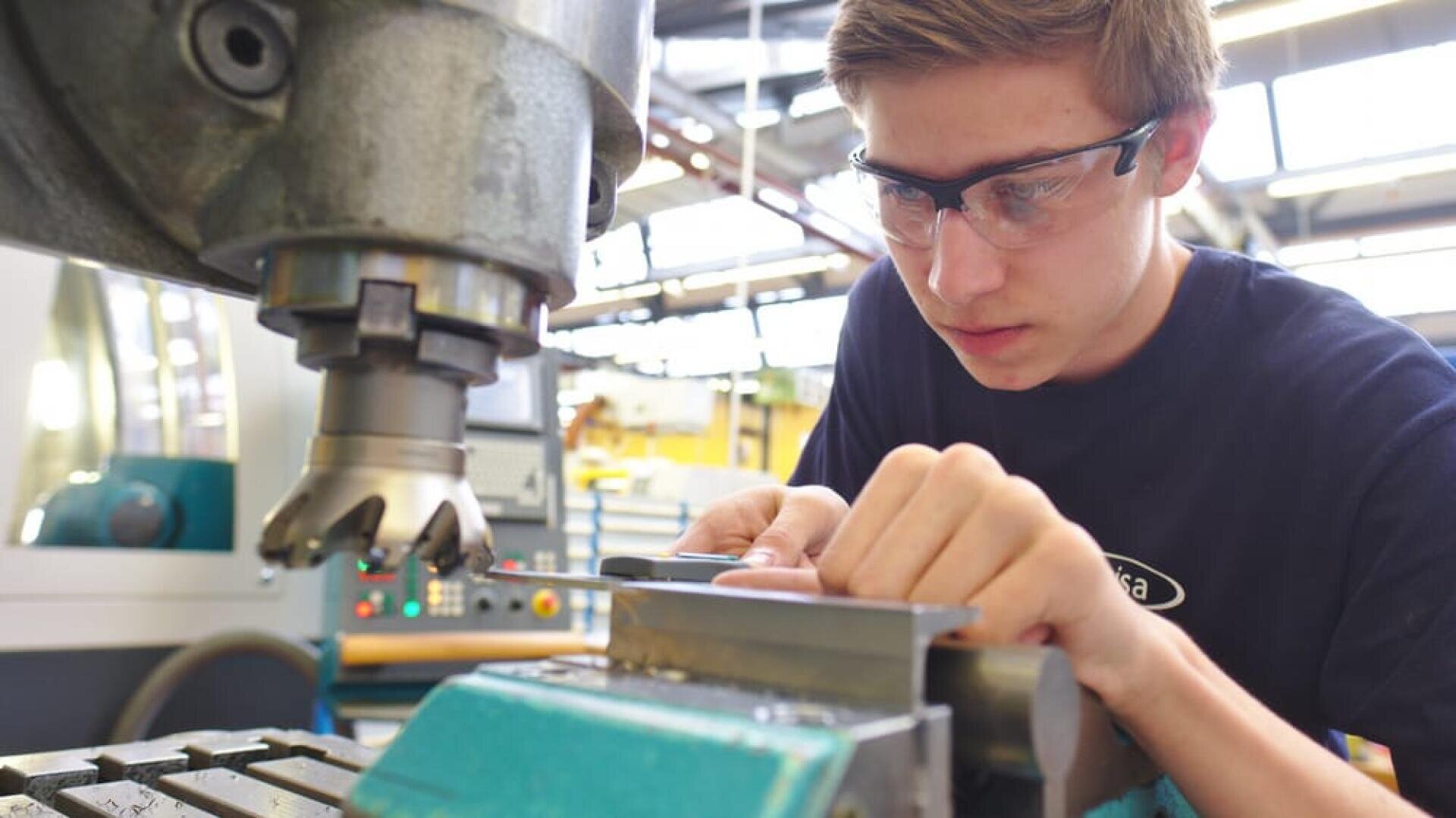 Ein junger Mann mit Schutzbrille bedient in einer Werkstatt eine Fräsmaschine und prüft das Metallstück, das er bearbeitet, genau.