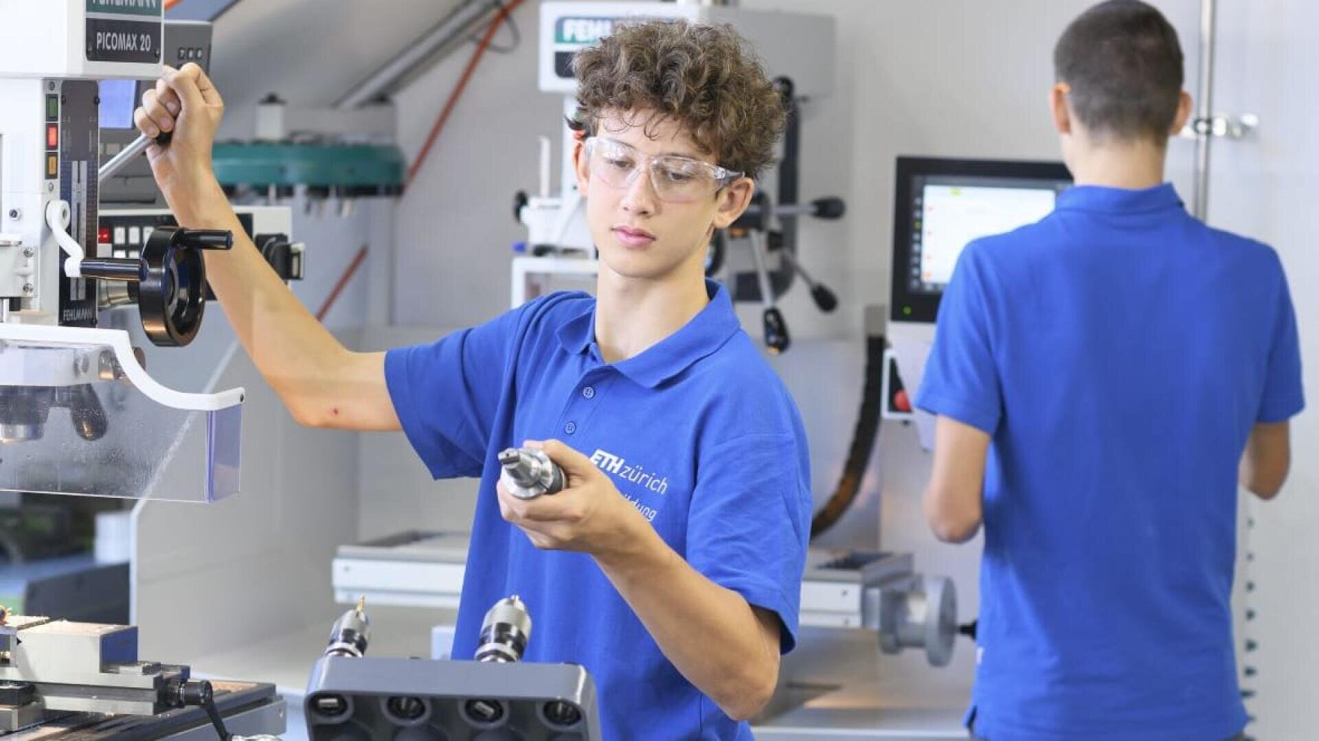 Ein junger Mensch mit Schutzbrille und blauem Poloshirt bedient in einer Werkstatt Maschinen und hält ein Werkzeug in der Hand, während im Hintergrund eine andere Person an einem Monitor steht.