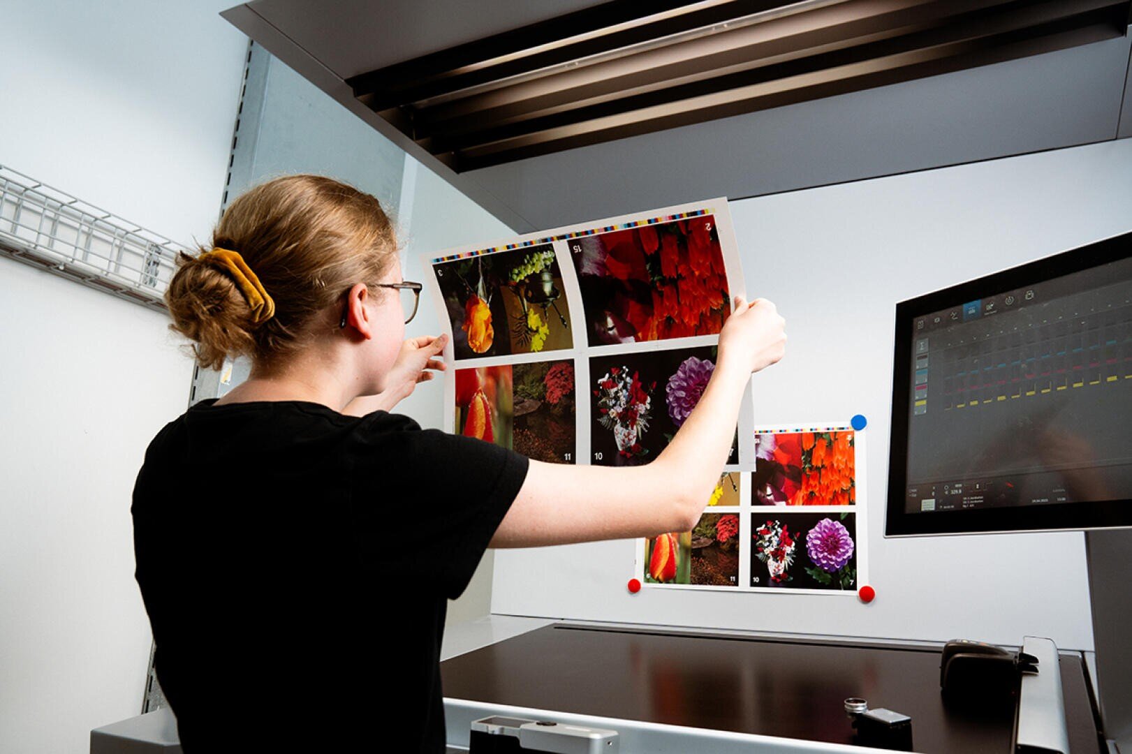 Eine Person mit zurückgebundenen Haaren hält einen Bogen mit bunten gedruckten Blumenbildern hoch und betrachtet sie unter einem Licht neben einem Monitor in einer Druckproduktion oder Qualitätskontrolle.