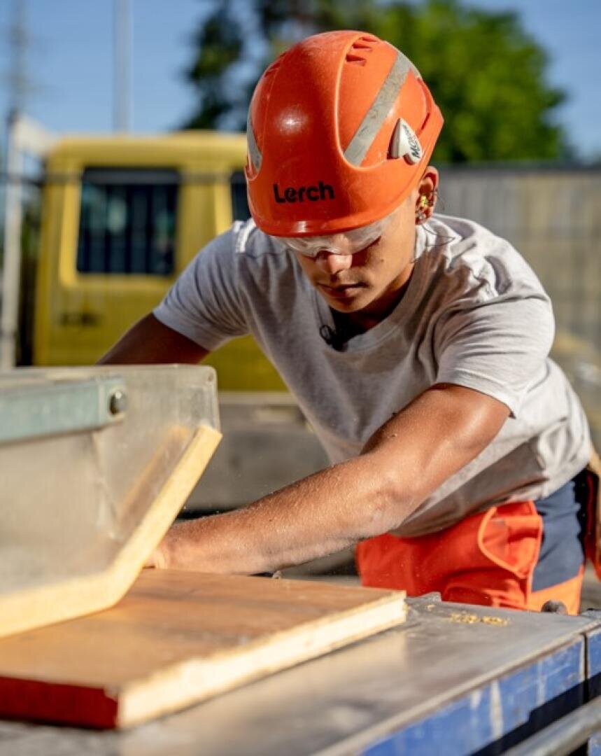 Ein Bauarbeiter, der einen orangefarbenen Schutzhelm und eine Schutzbrille trägt, bedient eine Tischsäge und sägt an einem sonnigen Tag im Freien ein Stück Holz. Im Hintergrund ist ein gelbes Fahrzeug zu sehen.