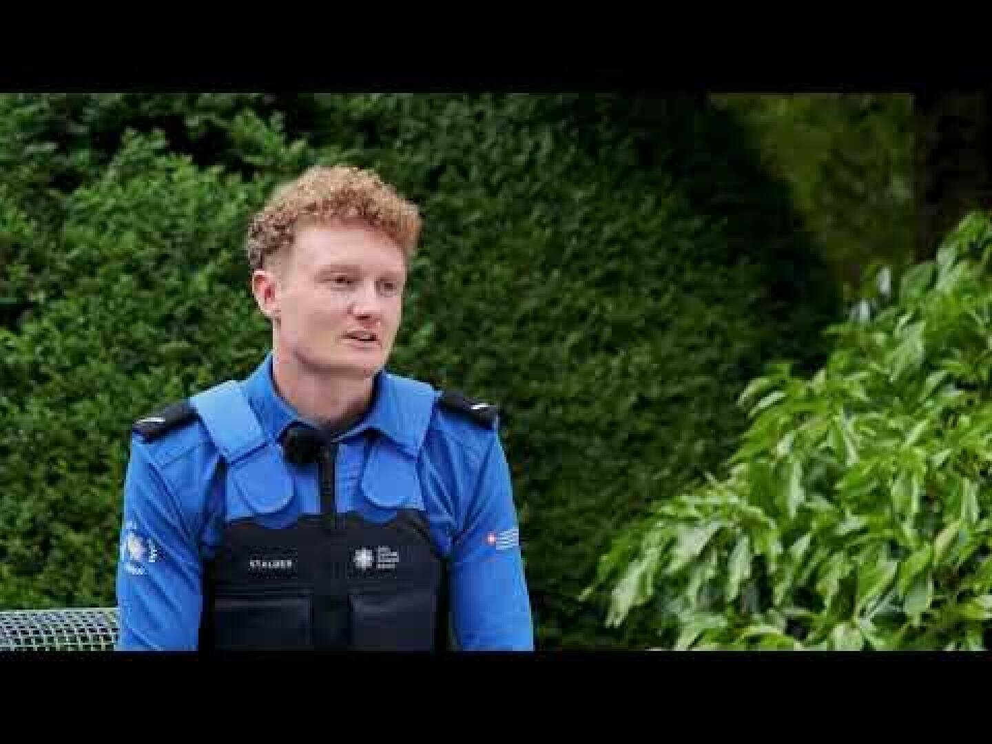 Eine Person in blauer Polizeiuniform sitzt im Freien auf einer Bank und spricht. Im Hintergrund sind üppige grüne Büsche und Pflanzen zu sehen.