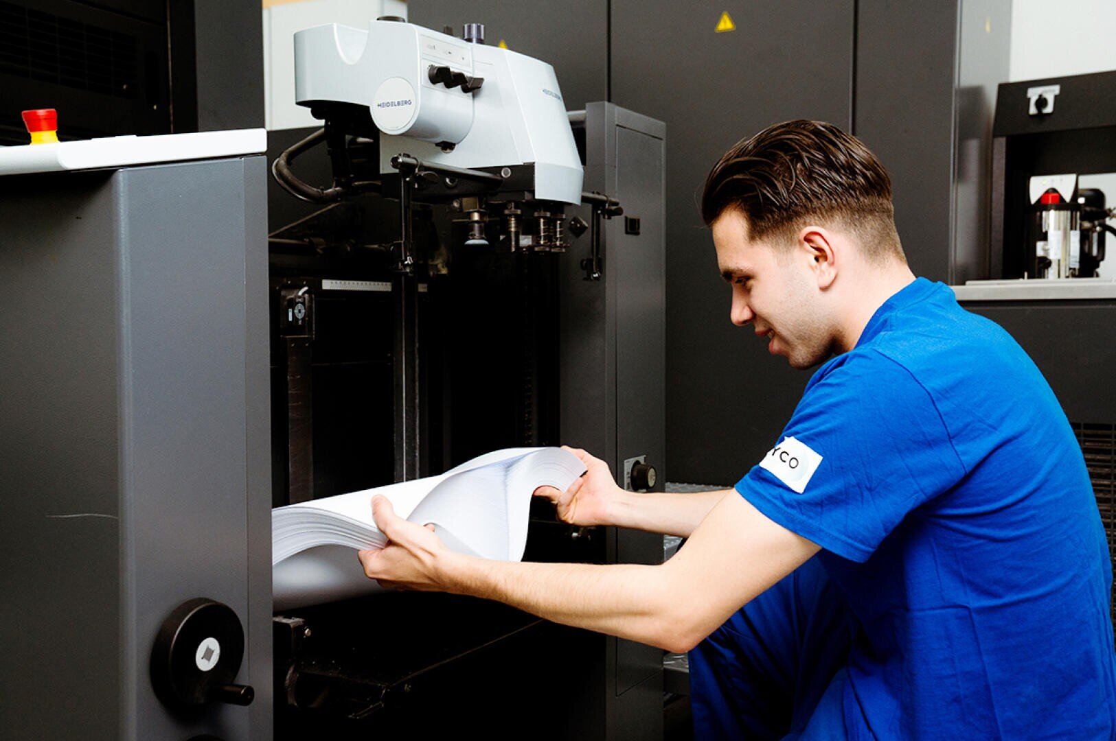 Ein Mann in blauer Arbeitskleidung bedient eine Druckmaschine und hantiert vorsichtig mit einem Stapel bedruckter Papiere, die aus dem Gerät in einer Industrieumgebung kommen.
