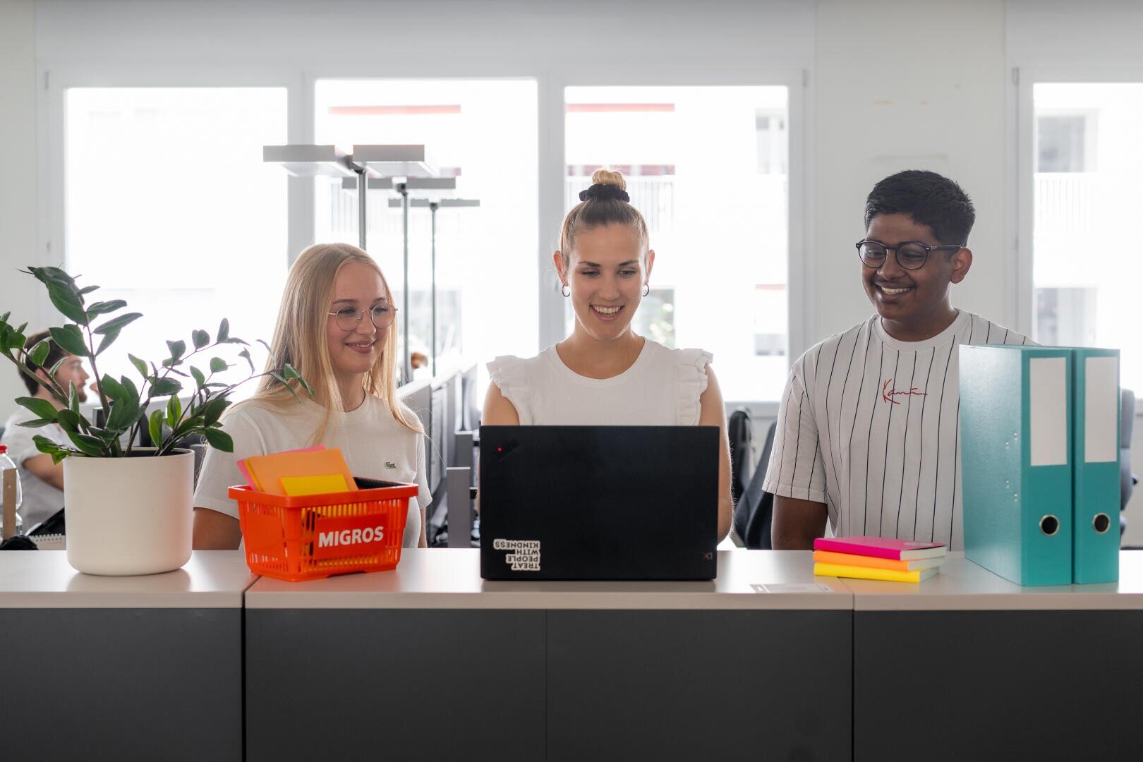 Drei junge Erwachsene stehen hinter einem Schreibtisch in einem Büro, lächeln und schauen auf einen Laptop. Auf dem Tresen stehen eine Topfpflanze, ein orangefarbener Korb, bunte Notizblöcke und zwei blaue Ordner. Im Hintergrund sind helle Fenster zu sehen.