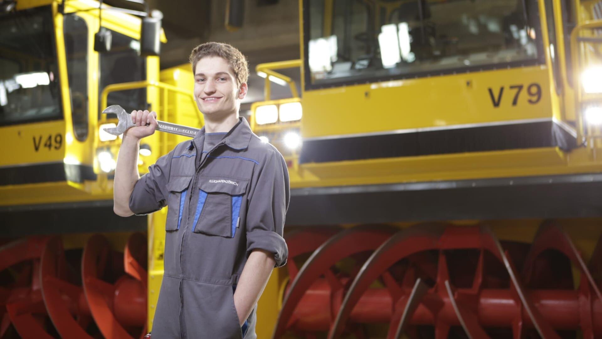 Ein junger Mann in einem grauen Arbeitsoverall hält einen großen Schraubenschlüssel über die Schulter und steht lächelnd vor einem leuchtend gelben Industriefahrzeug mit roten Rädern.