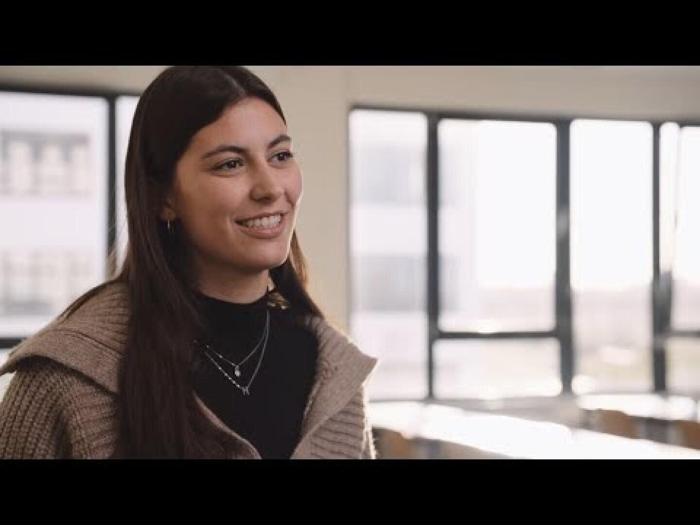 Eine junge Frau mit langen dunklen Haaren lächelt, während sie in einem hell beleuchteten Raum steht, im Hintergrund sind große Fenster zu sehen.