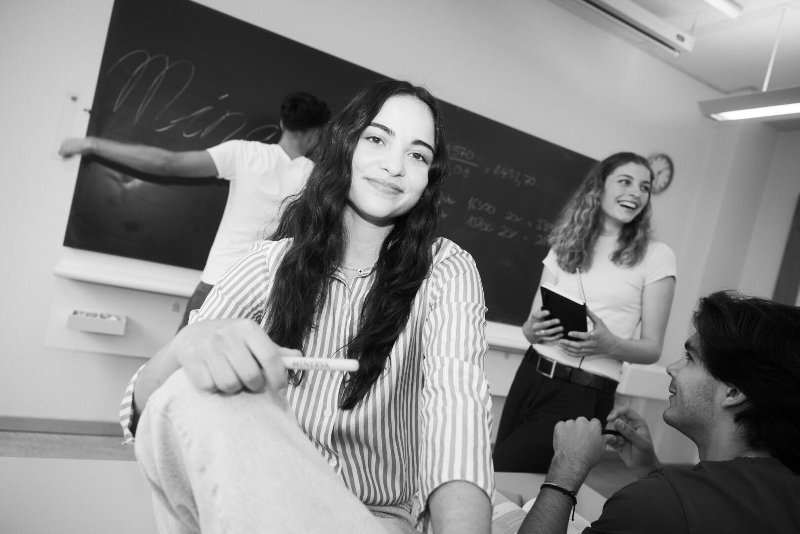 Eine junge Frau sitzt lächelnd im Vordergrund eines Klassenzimmers, während ein Mann an einer Tafel schreibt und zwei weitere Schüler im Hintergrund stehen und sich unterhalten. Die Szene ist freundlich und ungezwungen.