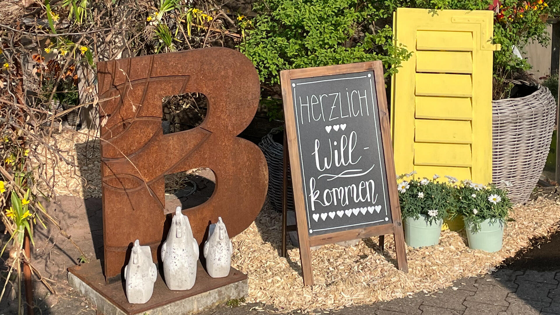 Ein großer Metallbuchstabe B, ein kleines Kreideschild mit der Aufschrift Herzlich Willkommen und Herzen, fünf dekorative weiße Flaschen und ein gelber Fensterladen sind zwischen Pflanzen und Blumen im Freien angeordnet.