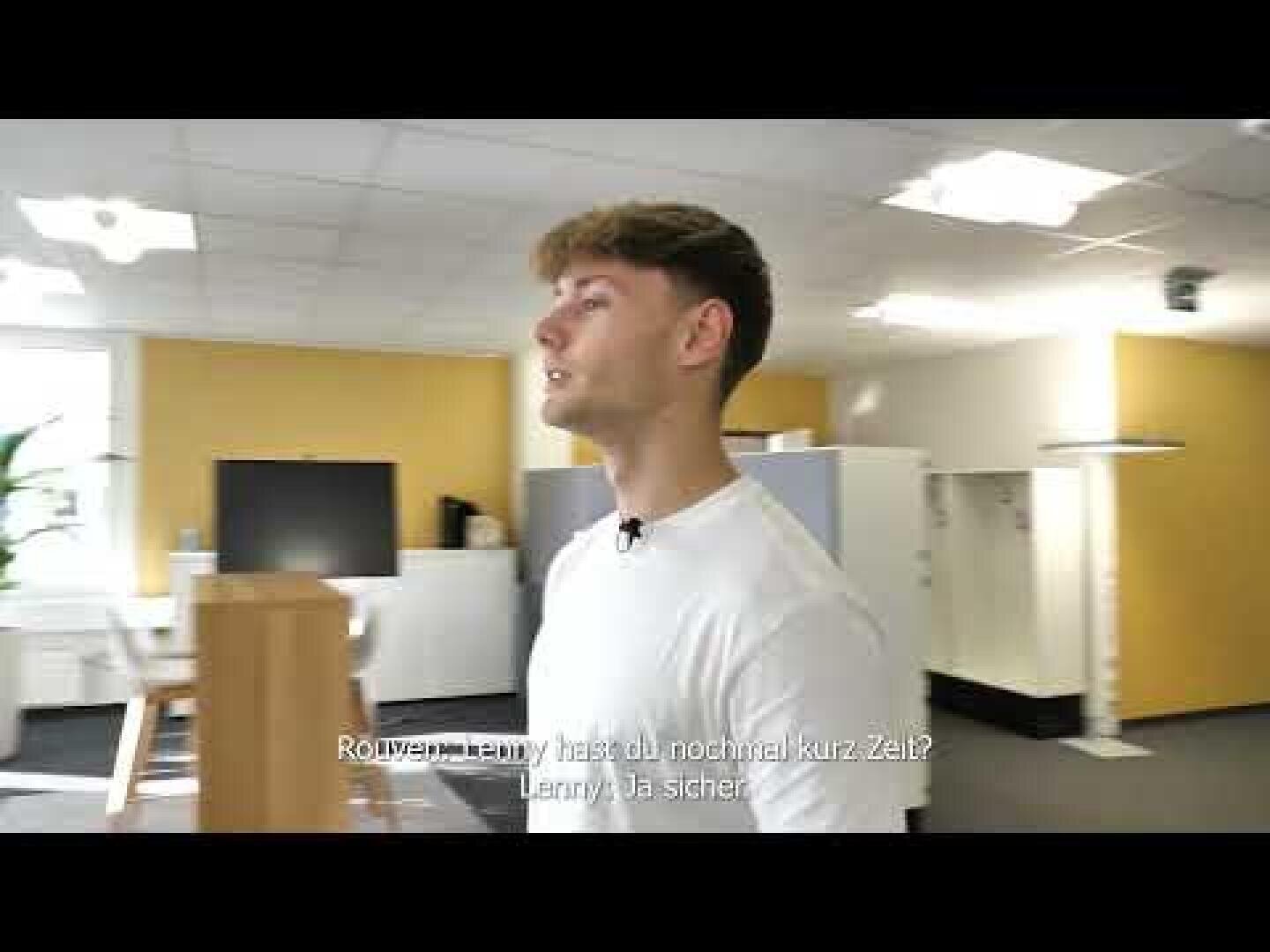 Ein junger Mann in einem weißen T-Shirt steht in einem modernen Büro mit gelben und weißen Wänden, Schränken und hellen Deckenleuchten. Niederländische Untertitel sind am unteren Rand des Bildes zu sehen.