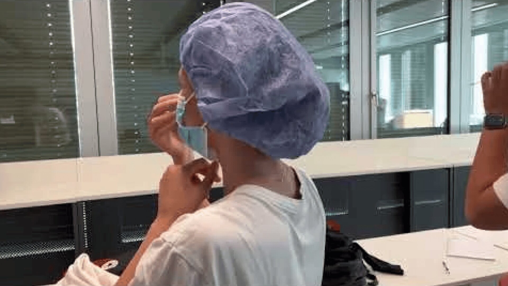 In einem hellen Raum mit großen Fenstern und moderner Einrichtung führt eine Person mit blauer OP-Mütze und weißem Hemd einen Tupfer in die Nase ein und führt wahrscheinlich einen COVID-19-Test durch.