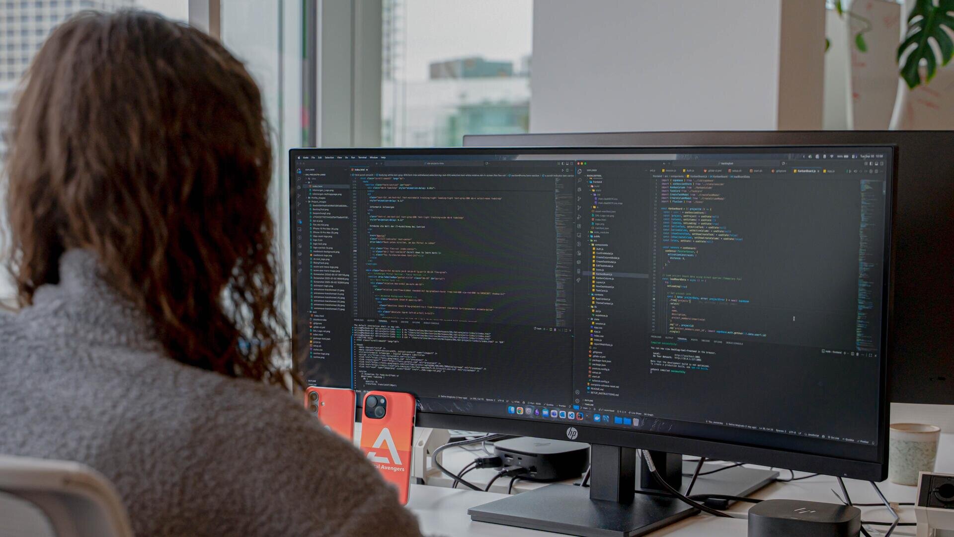Eine Person mit langen Haaren sitzt an einem Schreibtisch und betrachtet Codes auf einem großen Computermonitor in einem Büro mit großen Fenstern. Ein orangefarbenes Telefon und andere Büroutensilien stehen ebenfalls auf dem Schreibtisch.