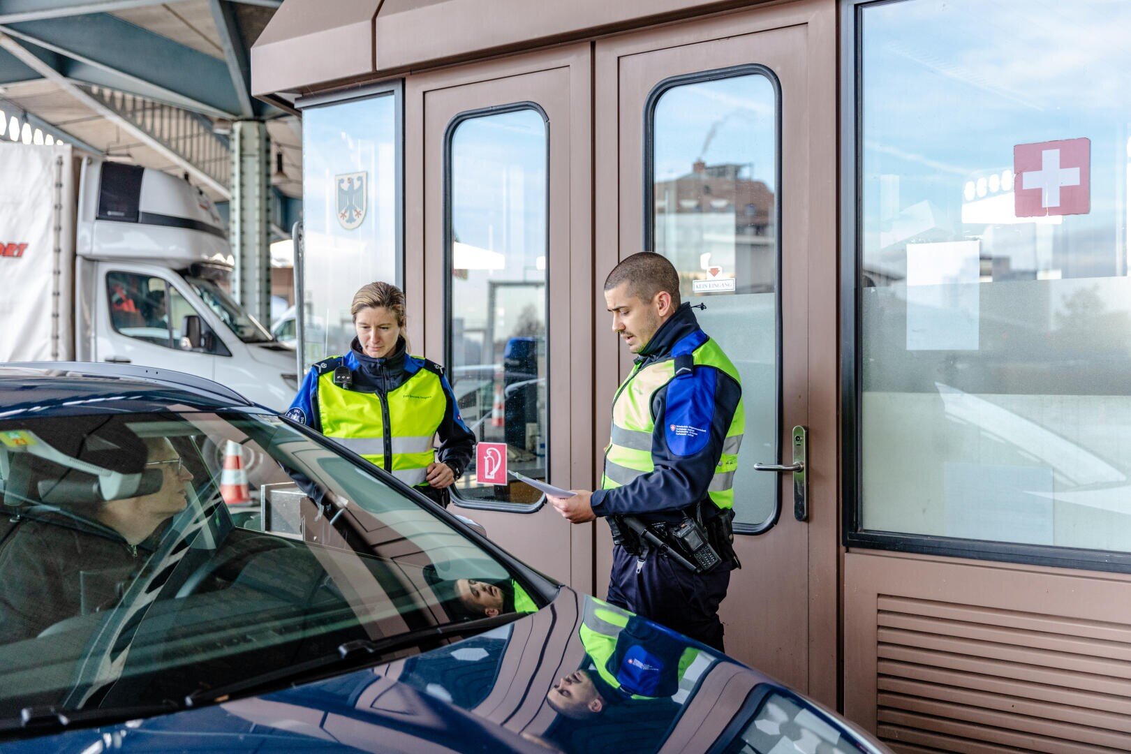 Zwei uniformierte Grenzbeamte stehen neben einem Auto und überprüfen die Dokumente des Fahrers an einem Grenzkontrollpunkt in der Nähe eines Gebäudes mit einem Schweizer Flaggensymbol am Fenster.