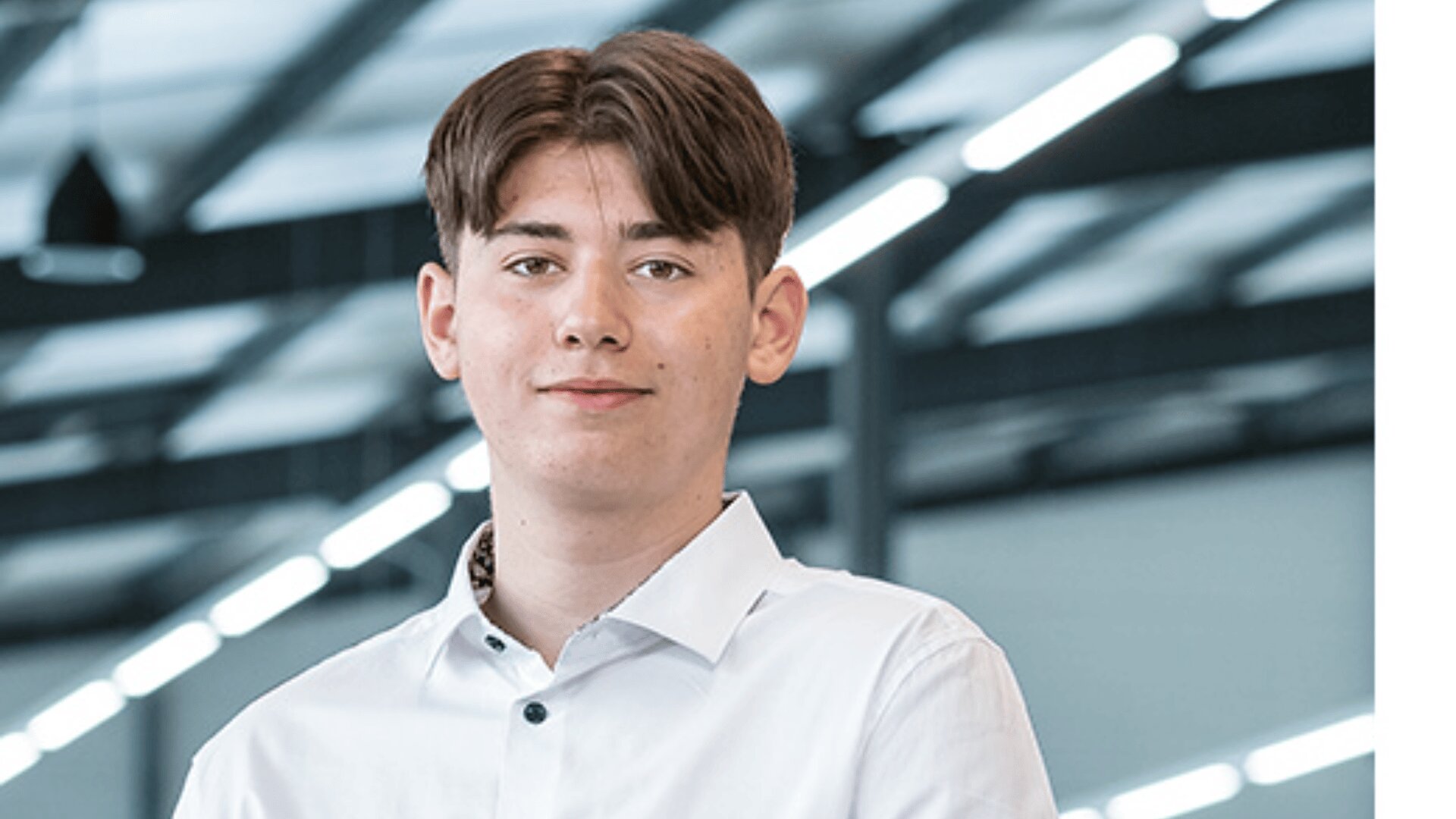 Ein junger Mann mit kurzen braunen Haaren und weißem Button-up-Hemd steht in einem Innenraum vor einem unscharfen Industriehintergrund mit hellen Oberlichtern.