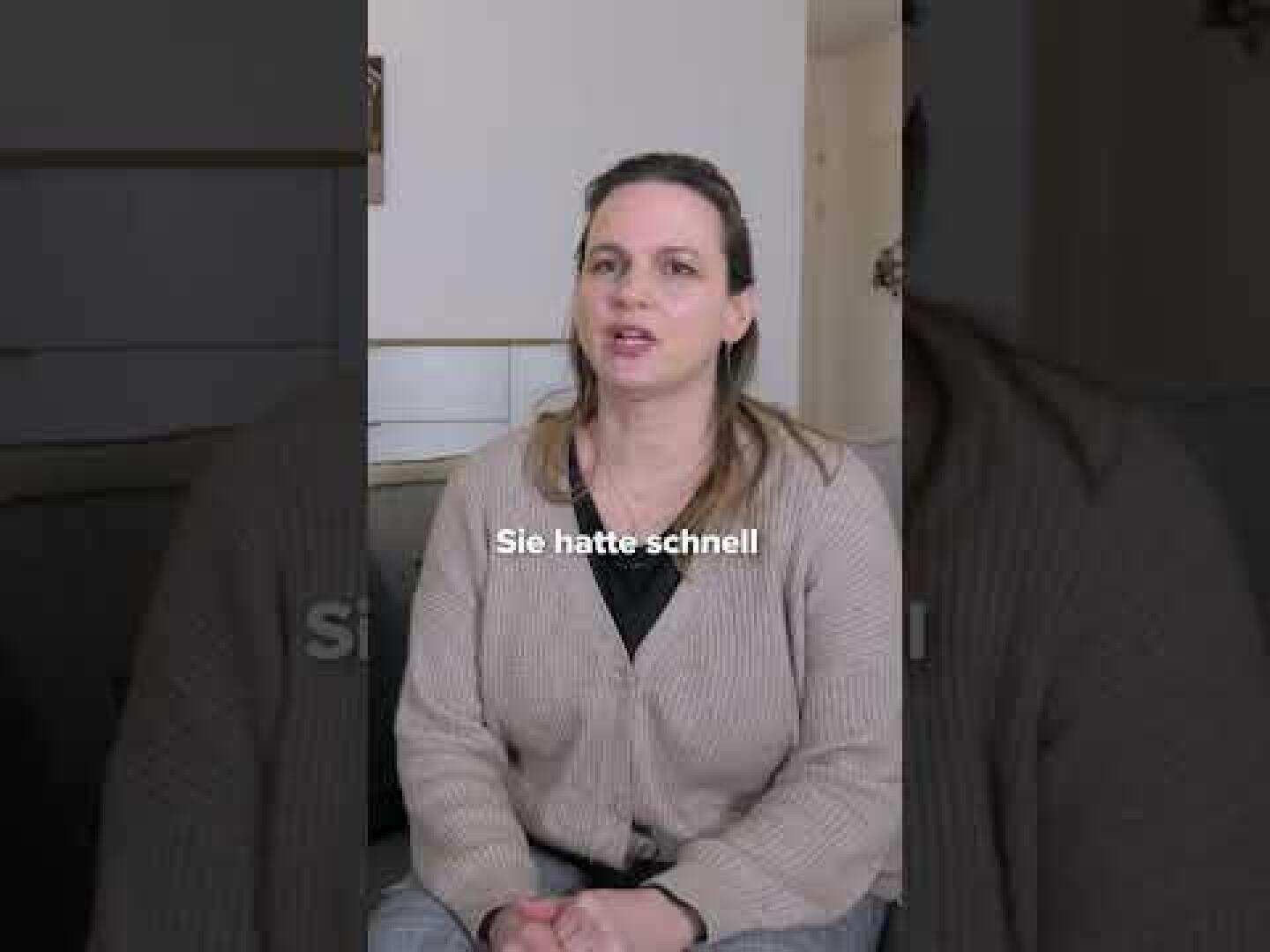 Eine Frau mit langen braunen Haaren sitzt in einem Raum und trägt eine beige Strickjacke und ein schwarzes Oberteil. Auf dem Bild ist der deutsche Text „Sie hatte schnell“ zu lesen. Im Hintergrund sind ein Teil eines Tisches und eine Wand zu sehen.