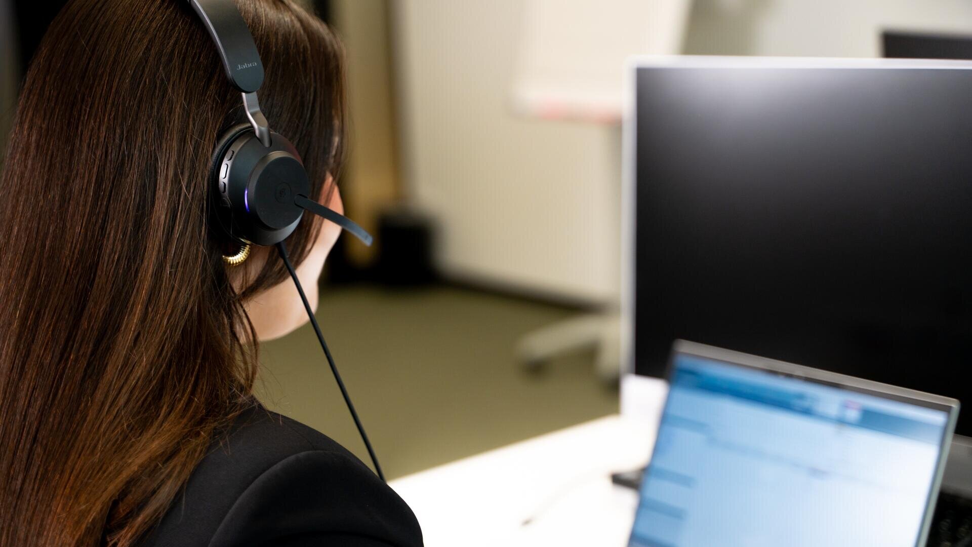 Eine Person mit langen braunen Haaren, die ein schwarzes Headset trägt, sitzt an einem Schreibtisch mit Blick auf einen Computermonitor und einen Laptop mit unscharfem Bildschirm in einer Büroumgebung.