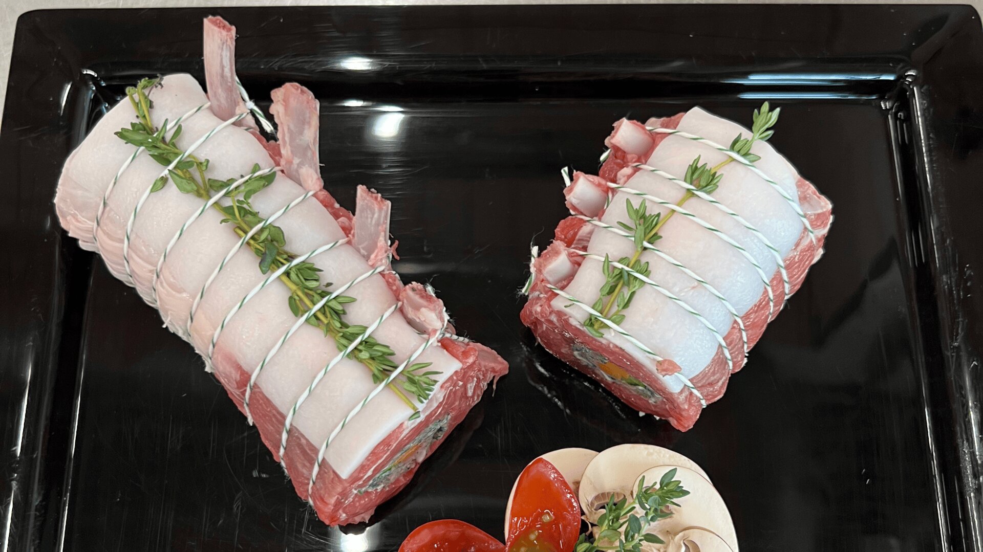 Zwei rohe Lammkarrees, mit Schnur gebunden und mit Rosmarinzweigen garniert, auf einem schwarzen Tablett platziert. In Scheiben geschnittene Tomaten und Champignons liegen als Garnitur daneben.