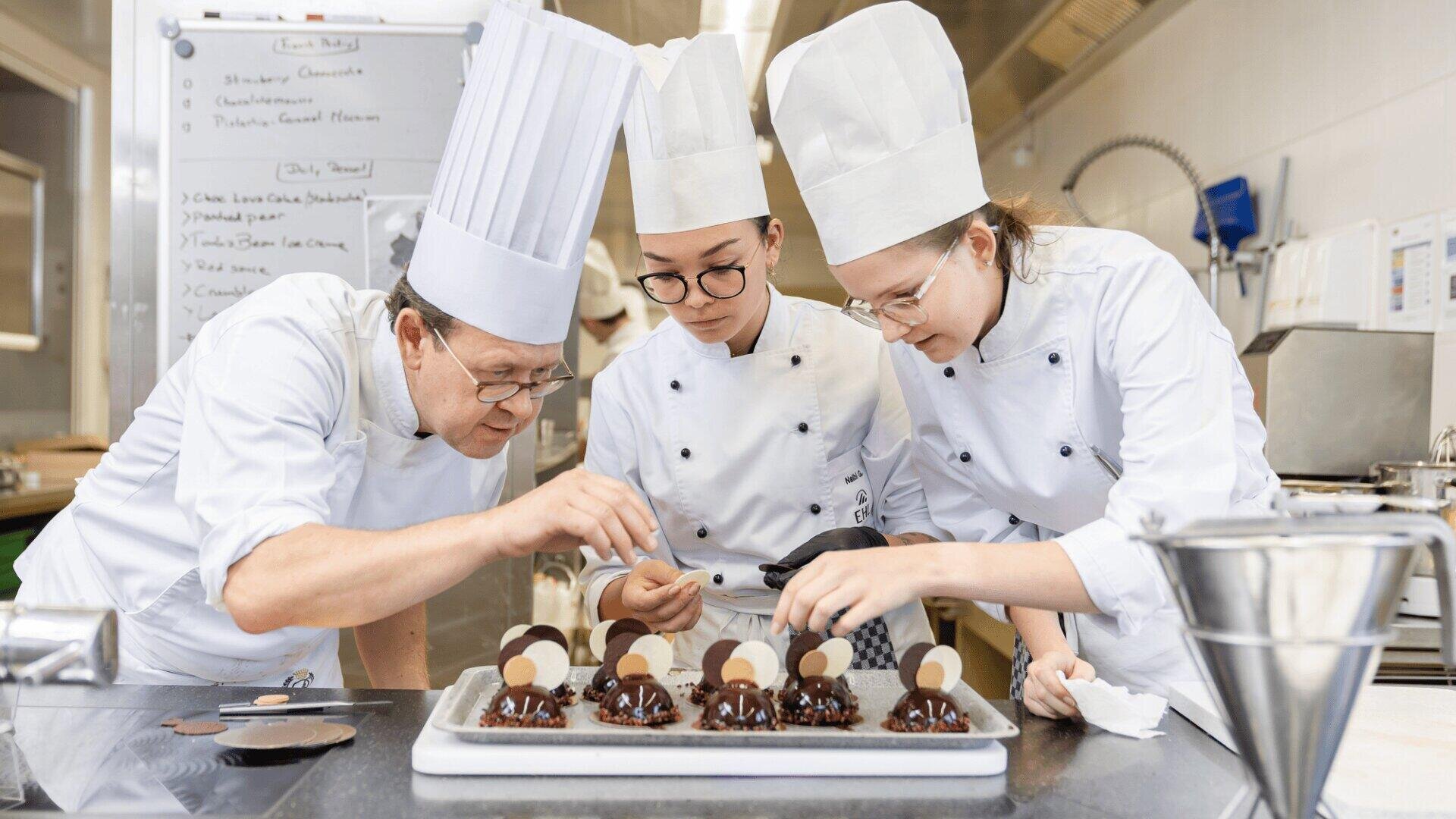 Drei Konditoren in weißen Uniformen und Mützen dekorieren in einer Profiküche sorgfältig Schokoladendesserts auf einem Tablett, im Hintergrund sind eine Weißwandtafel und Küchenutensilien zu sehen.