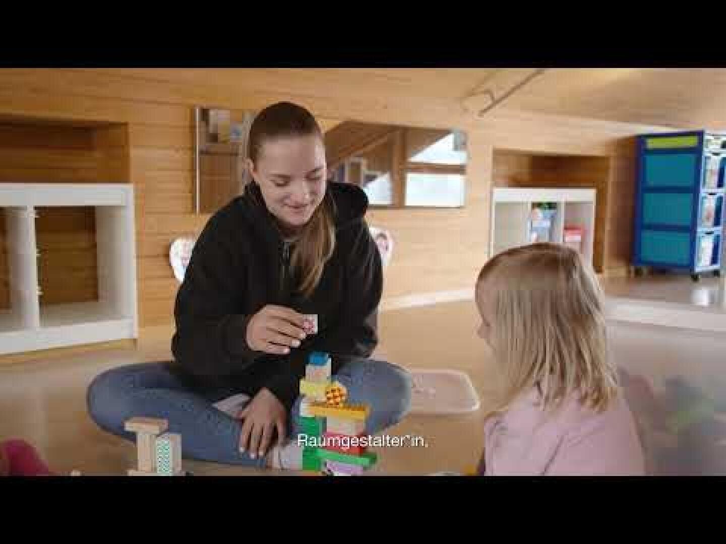 Zwei Personen, ein Erwachsener und ein Kind, sitzen in einem warm beleuchteten Raum mit Holzwänden auf dem Boden und spielen mit bunten Bauklötzen. Der Erwachsene lächelt und hält einen Klotz in der Hand. Die Untertitel lauten Raumgestalter:in.