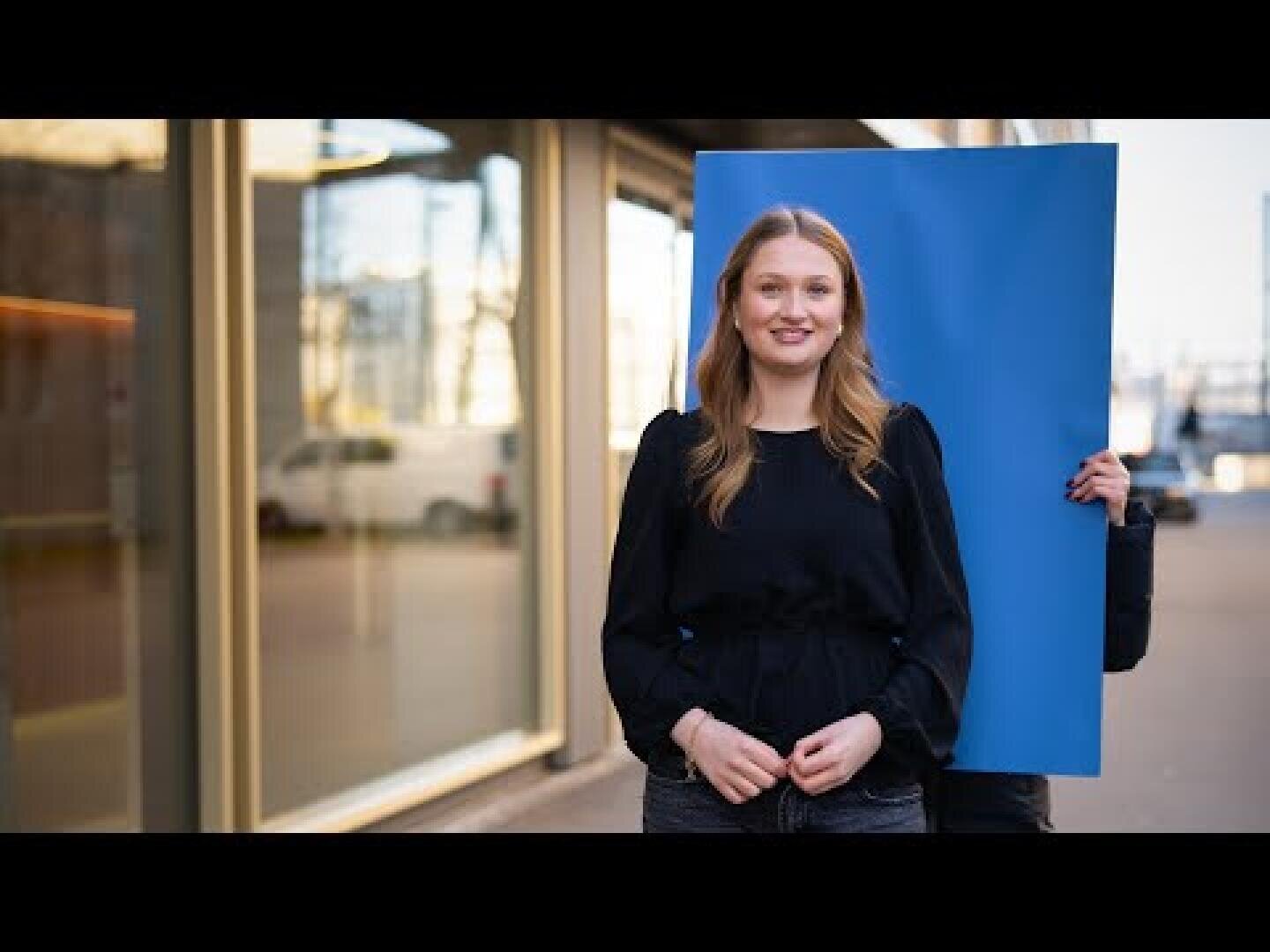Eine Frau steht draußen vor einem Glasgebäude und lächelt, während eine andere Person hinter ihr eine große blaue Plakatwand hochhält und sich selbst teilweise verdeckt.