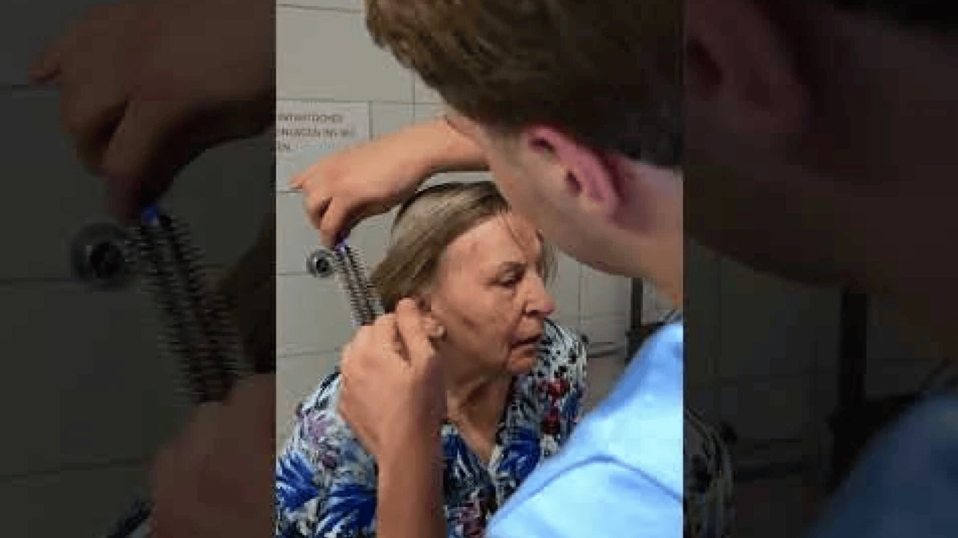 Eine Person hilft einer älteren Frau, ihr Haar aus einer großen Metallfeder zu entwirren, die in der Nähe ihres Kopfes steckt. Die Frau wirkt verzweifelt, und sie befinden sich in einem gekachelten Raum.