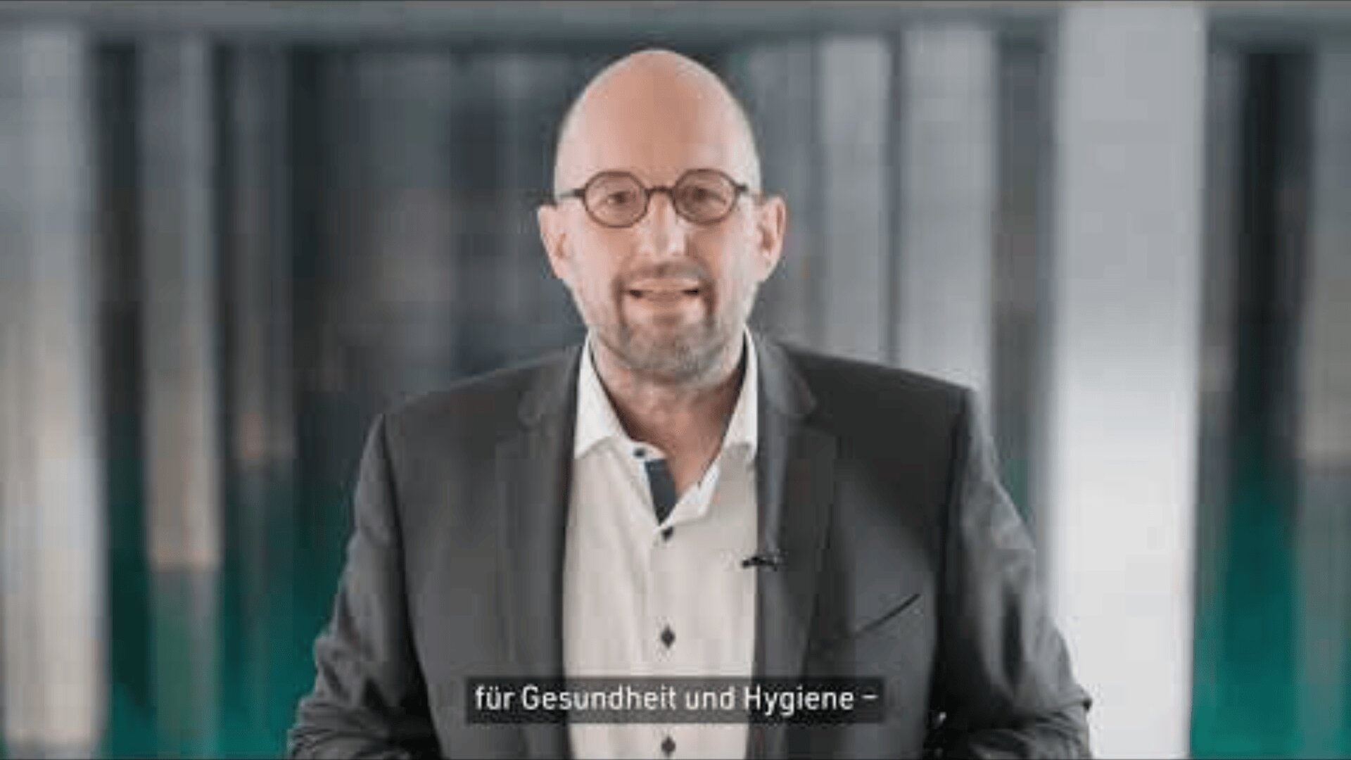 Ein glatzköpfiger Mann mit Brille und Bart, der einen dunklen Anzug und ein weißes Hemd trägt, steht vor einem unscharfen Hintergrund. Die deutschen Untertitel am unteren Rand lauten "für Gesundheit und Hygiene -".
