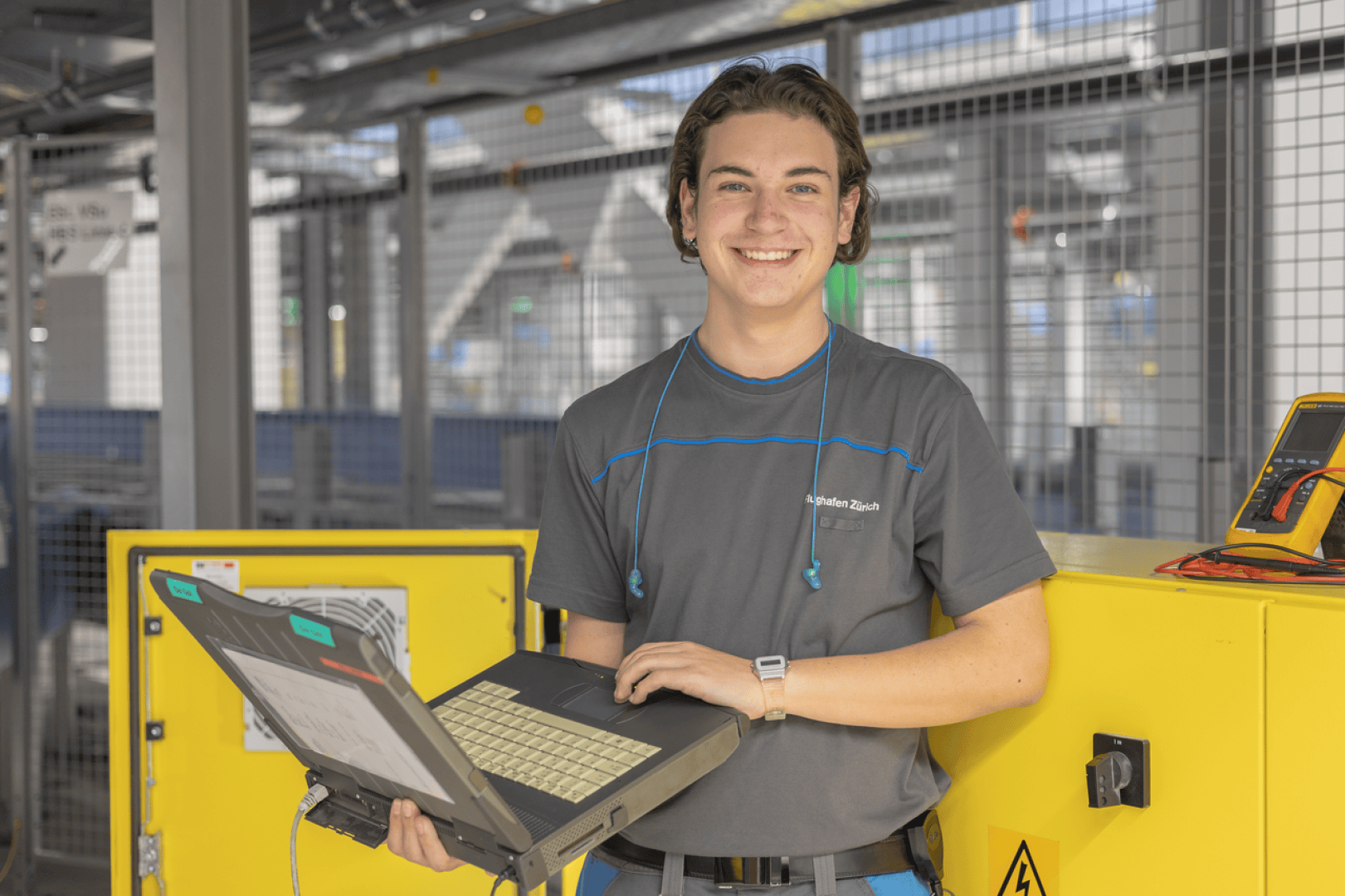 Ein lächelnder junger Techniker in einer grauen Uniform steht mit einem aufgeklappten Laptop in einer Industrieumgebung mit gelben Schalttafeln und Geräten im Hintergrund.