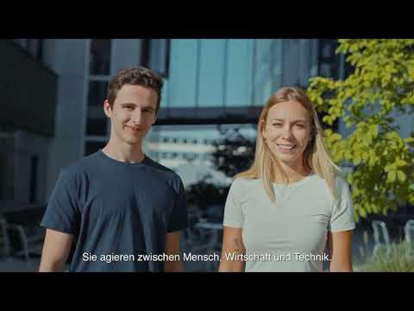 Ein junger Mann und eine junge Frau stehen draußen in einem sonnigen Innenhof und lächeln in die Kamera. Im Hintergrund sind Gebäude und Bäume zu sehen. Am unteren Bildrand erscheint ein deutscher Text.