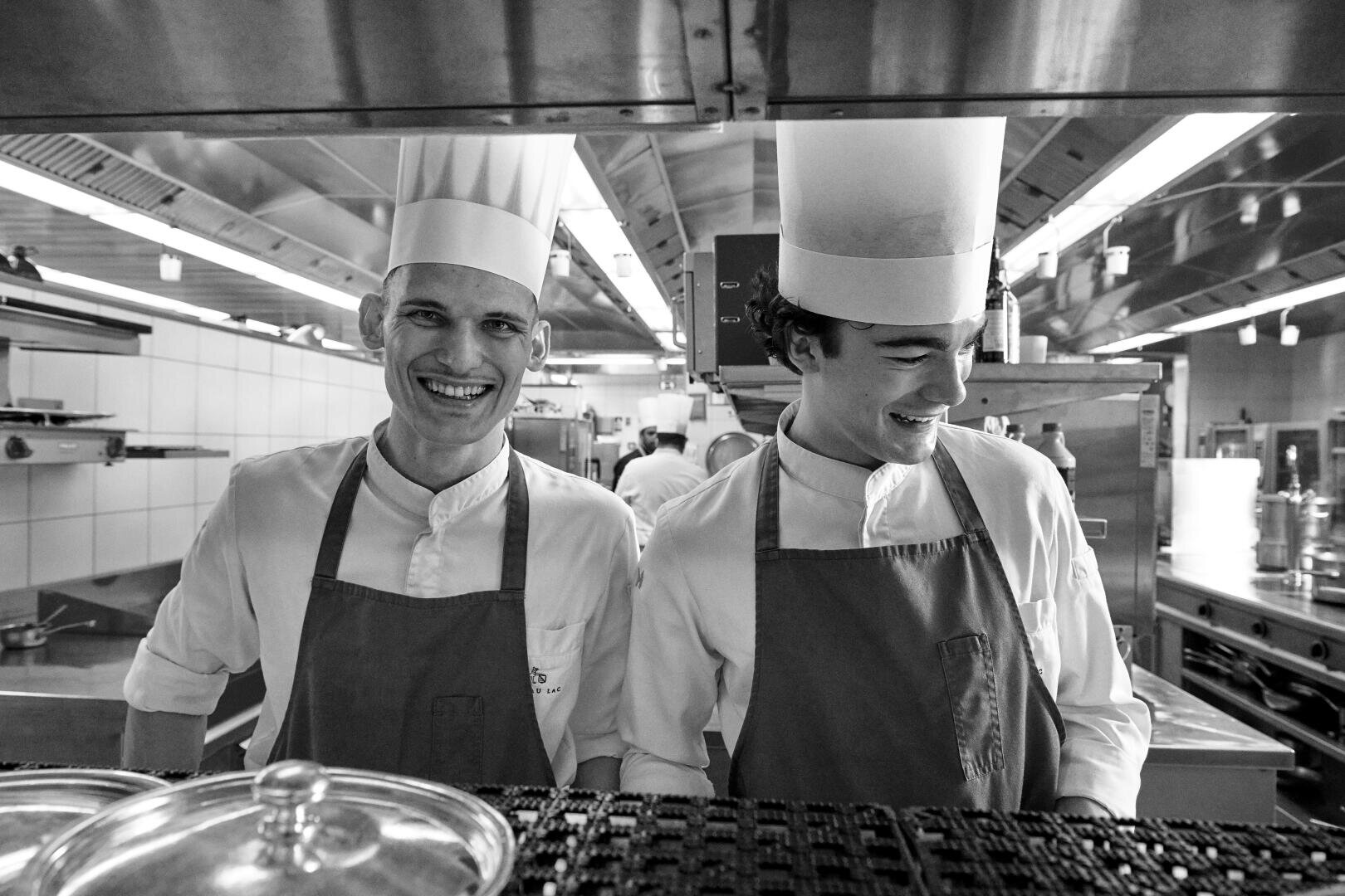 Zwei Köche in weißen Uniformen und hohen Mützen stehen Seite an Seite in einer professionellen Küche und lächeln und lachen zusammen. Im Hintergrund sind Küchengeräte und Regale zu sehen. Das Bild ist in schwarz-weiß gehalten.
