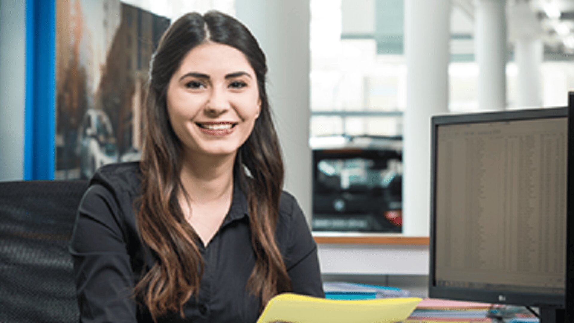 Eine Frau mit langen dunklen Haaren und schwarzem Hemd lächelt, während sie in einem Büro an einem Schreibtisch mit Computermonitor sitzt und gelbe Papiere hält.