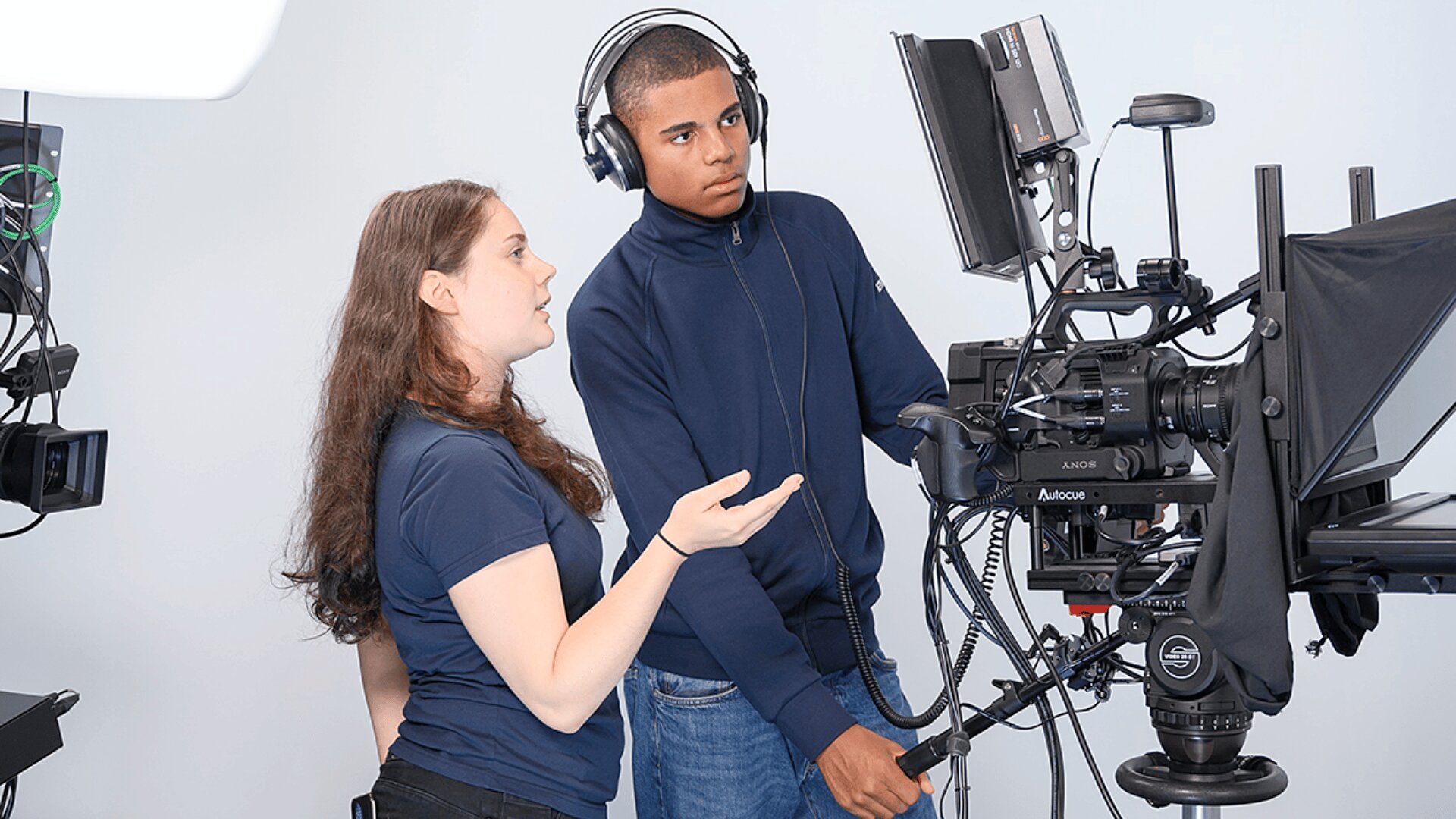 Zwei junge Leute bedienen eine professionelle Filmausrüstung; der eine trägt Kopfhörer und stellt die Kamera ein, während der andere mit ihm in einer Studioumgebung gestikuliert und etwas bespricht.