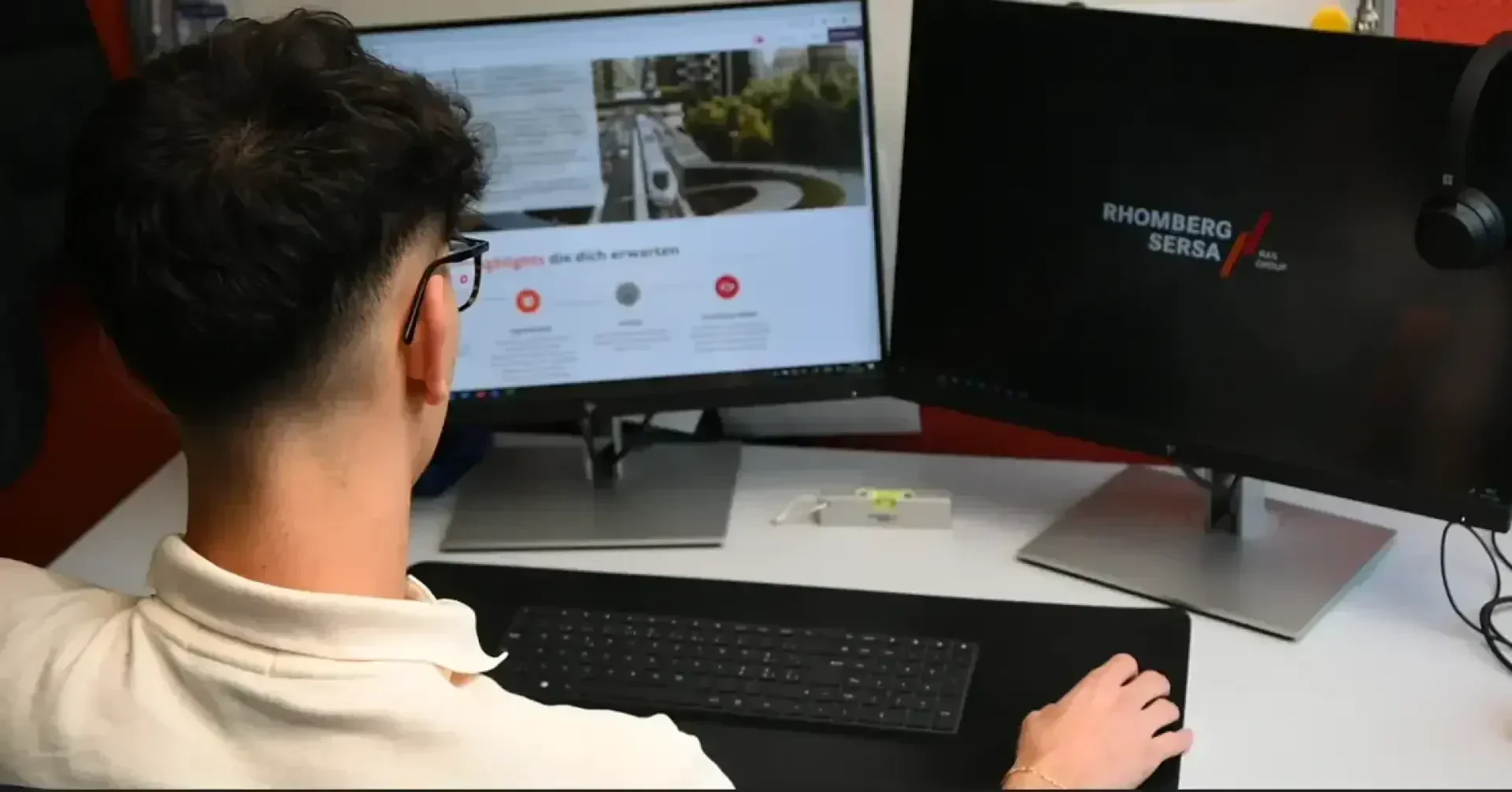 Eine Person mit kurzen, dunklen Haaren und einer Brille sitzt an einem Schreibtisch und arbeitet an einem Computer mit zwei Bildschirmen. Auf einem Bildschirm ist eine Website zu sehen, auf dem anderen das Logo von Rhomberg Sersa. Auf dem Schreibtisch liegen eine Tastatur und eine Maus.